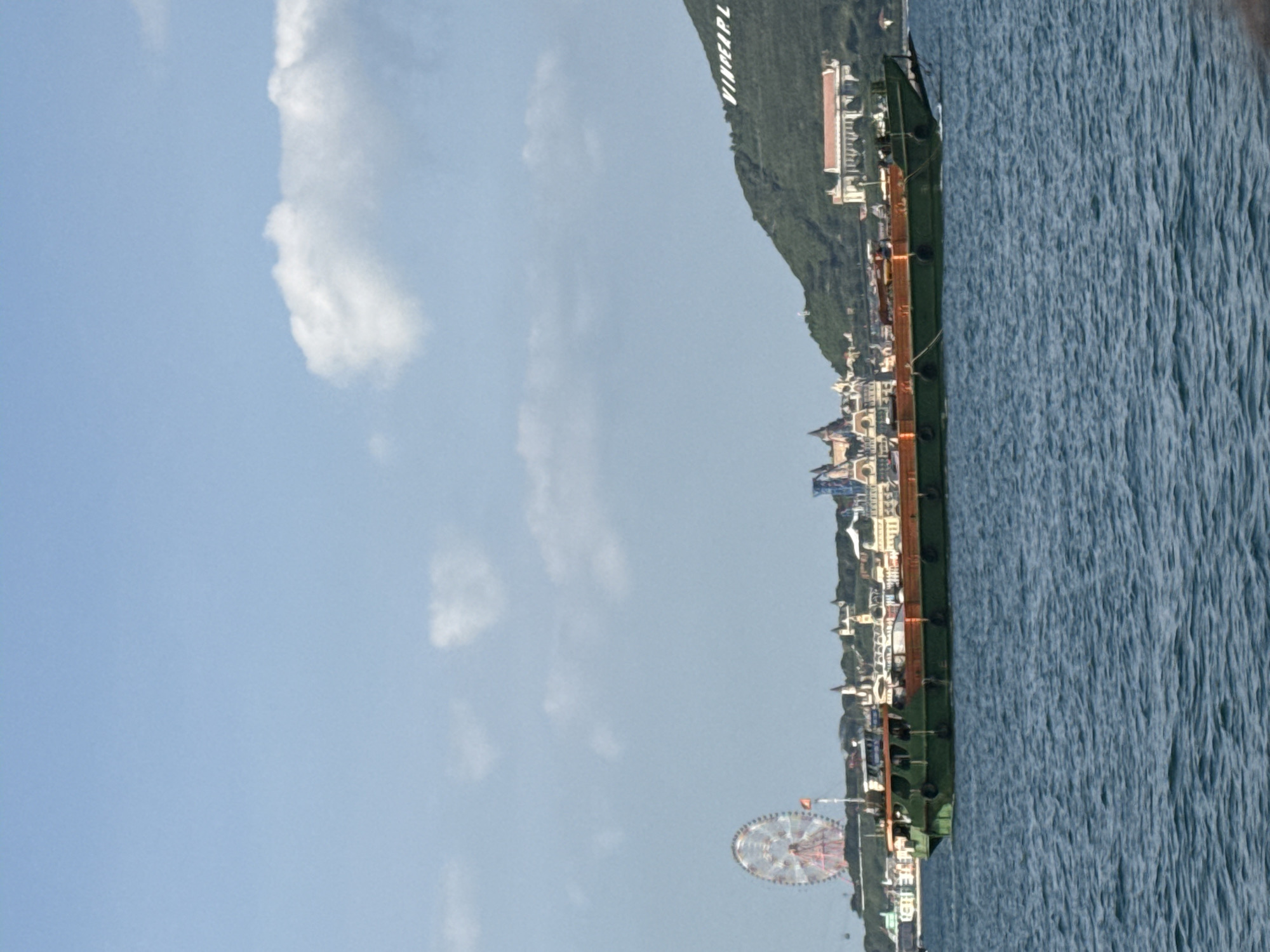 A green cargo barge travels on the water in front of the coast of Nha Trang Ward, Vietnam. A Ferris wheel and various buildings are visible on the coastline, with a large hill in the background. The sky is blue with some white clouds.