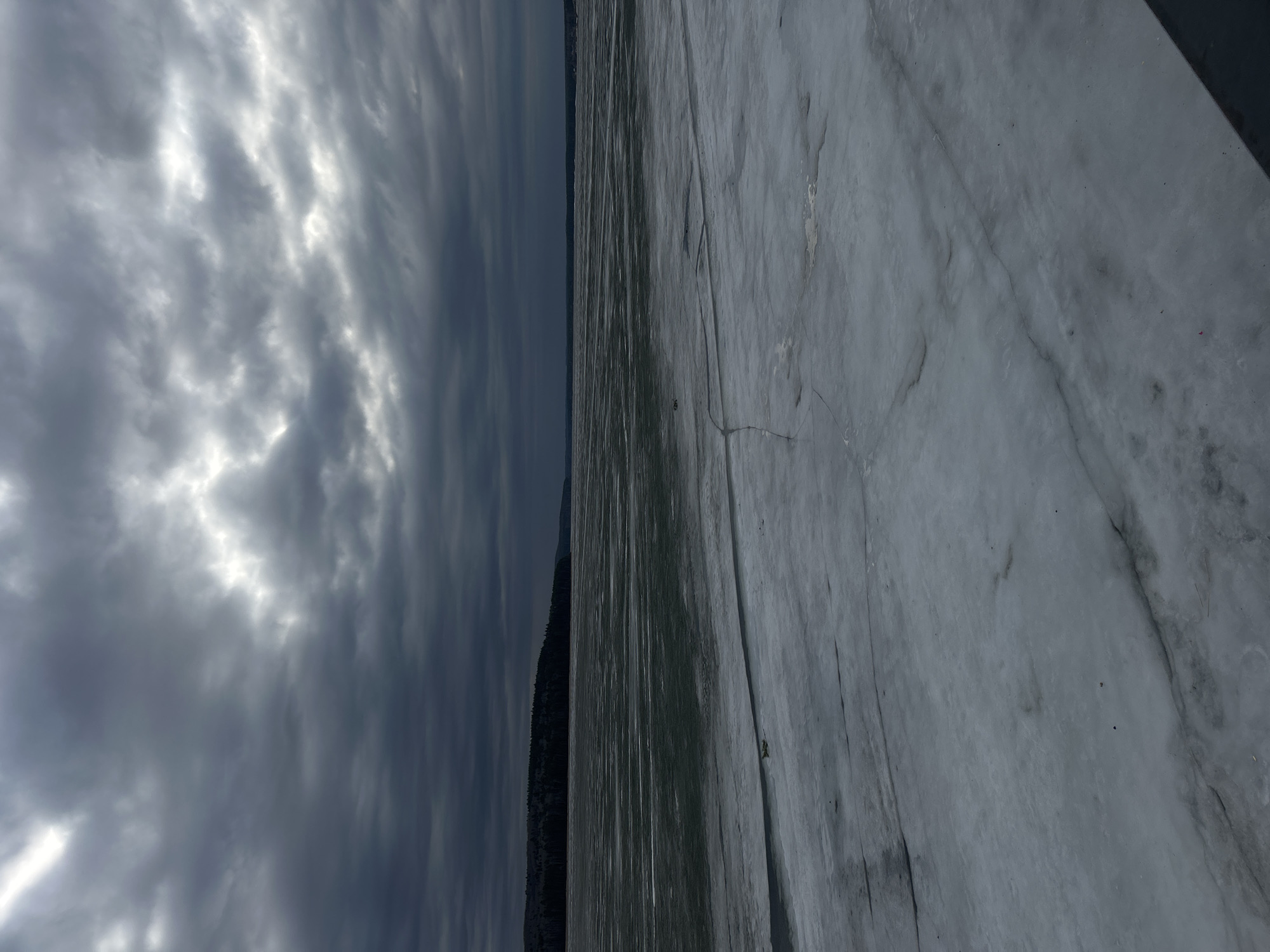 The image depicts a frozen lake or river with a cloudy sky overhead. A line of trees can be seen on the opposite shore. The surface of the ice is textured with cracks and patterns. Given the prompt, the location is likely in or near Chaikovsky, Russia. No people or animals are clearly visible, though there may be a distant figure on the ice.