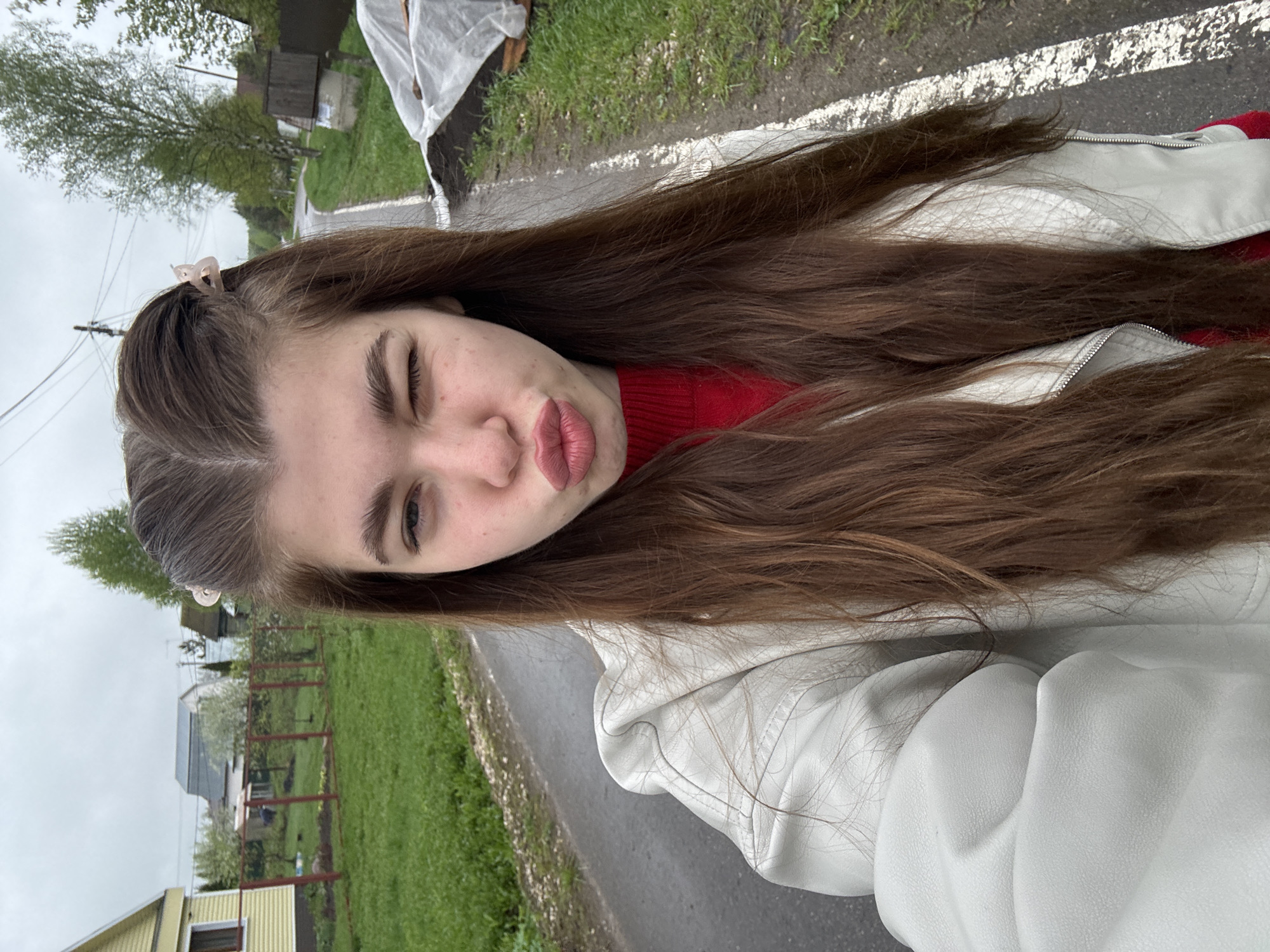 A young woman stands outdoors on a grey asphalt road, possibly in a residential area based on the background. She is wearing a red sweater and a white leather jacket, with long brown hair. A hair clip adorns her hair on the left side of the frame. She is winking with her left eye and pouting. The background features grass, a metal fence, trees, and houses. The sky appears overcast. It is Meshera, Russia.