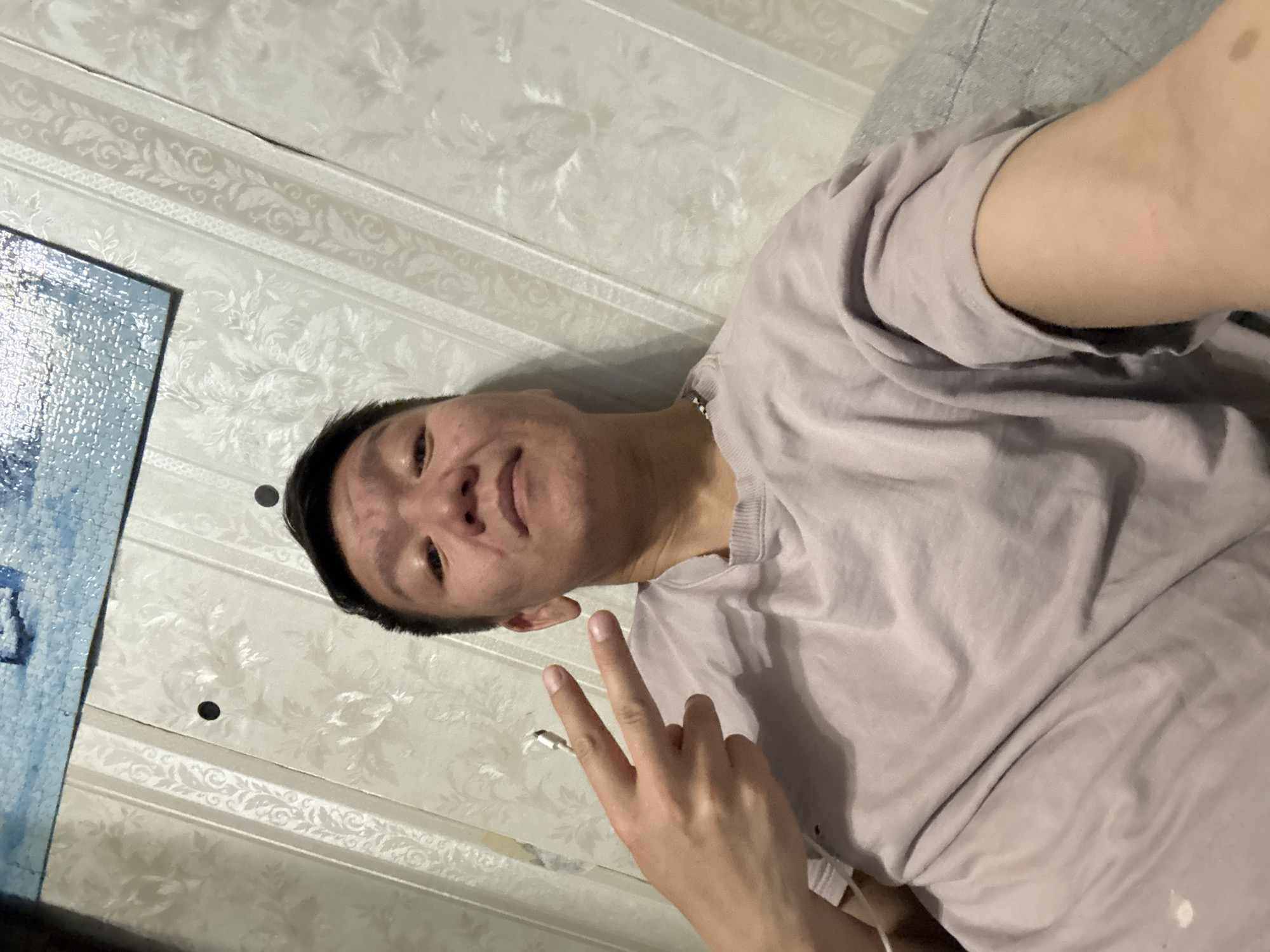A young man is pictured indoors, sitting on a couch. He is looking towards the camera and holding up two fingers in a peace sign with his right hand. He is wearing a light purple t-shirt. The background consists of a wall with floral patterned wallpaper and what appears to be a puzzle on the wall above. The image was taken in Khimki, Russia.