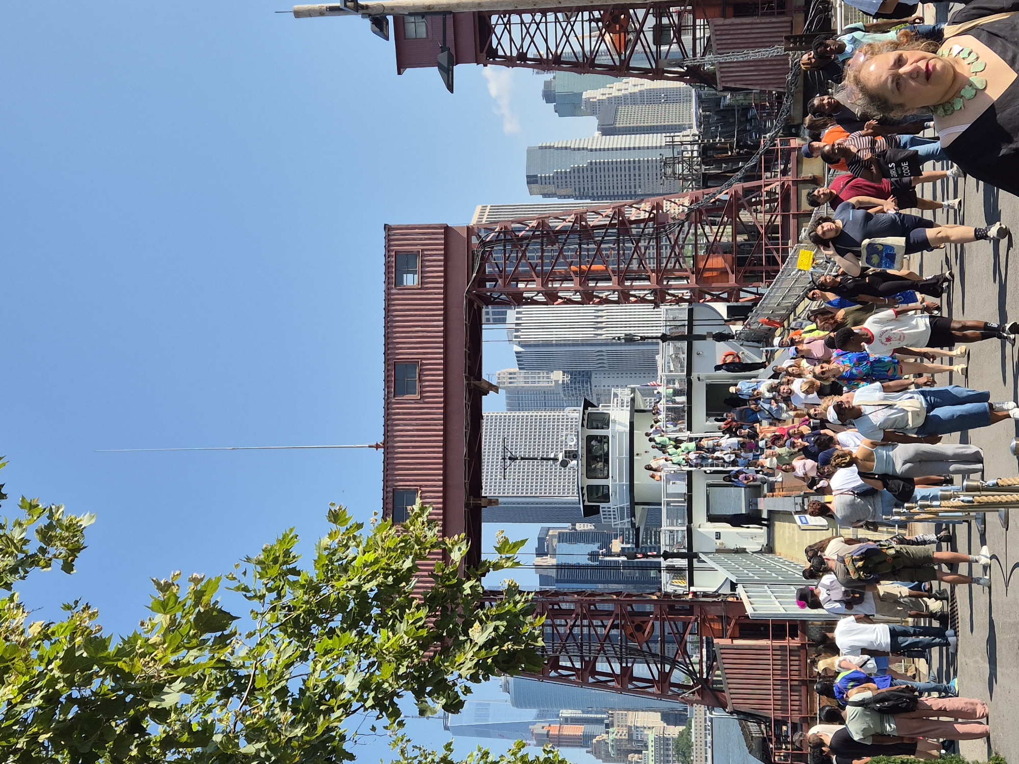 The image captures a bustling scene at a ferry terminal in New York City, likely part of the Staten Island Ferry system, given the characteristic red gantries and the distant view of the Manhattan skyline. A large crowd of people are gathered on the pedestrian walkway, preparing to board a ferry. The ferry itself is white with a dark hull and appears to be docked at a pier. Many individuals are walking towards the ferry, while others are already on its deck, ascending a ramp. The background is dominated by the iconic skyscrapers of Lower Manhattan, including One World Trade Center. The weather is clear and sunny, with a bright blue sky. The overall impression is one of activity and movement, with a significant number of tourists and commuters utilizing the ferry service.