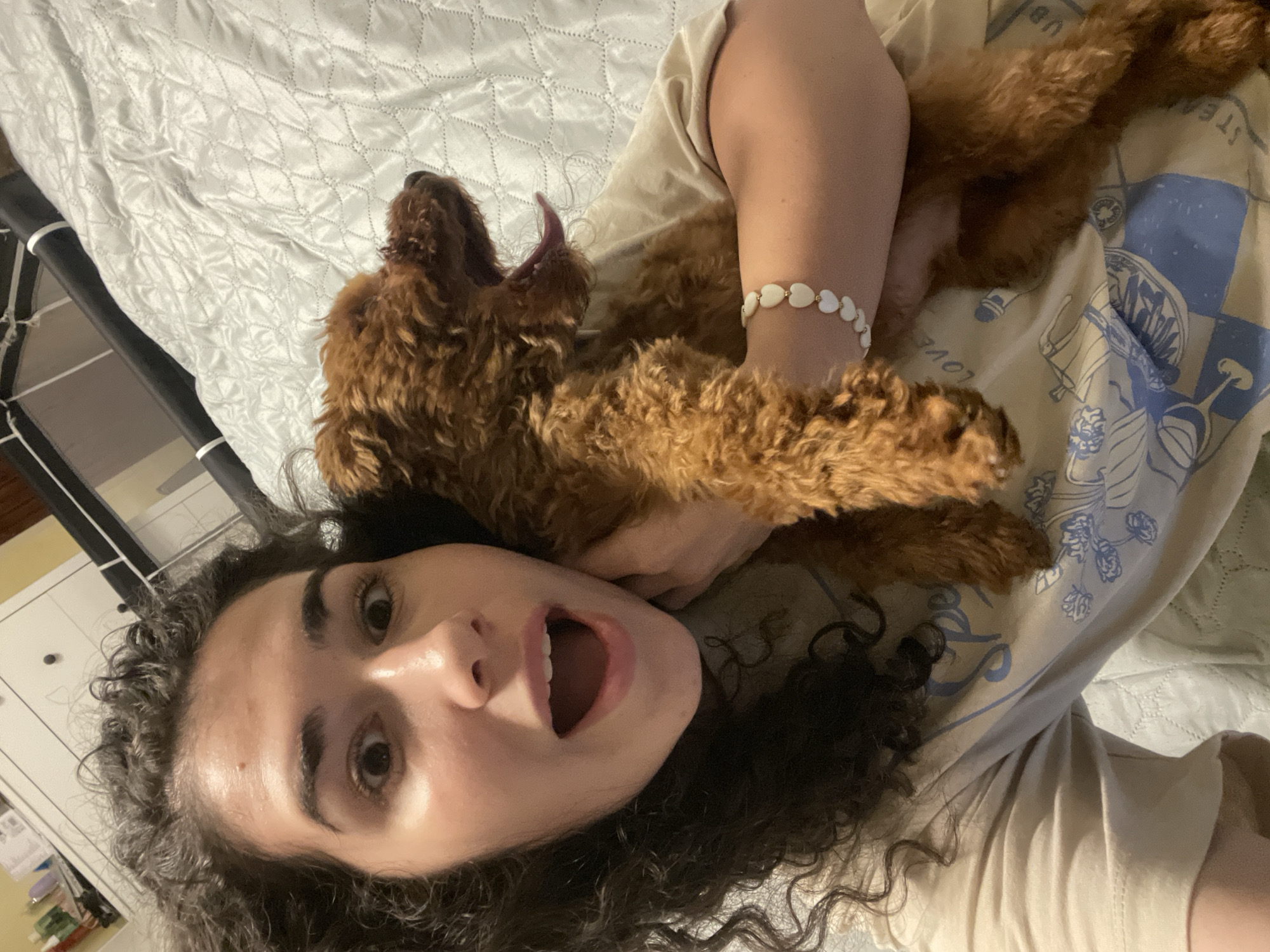 A person with dark, curly hair is lying on a bed with a light gray patterned duvet cover, holding a small, brown, curly-haired dog. The person is looking at the camera with their mouth open in an expression of surprise or excitement. The dog has its mouth open as well, appearing to yawn or pant. The person is wearing a light-colored t-shirt with a faint design. A white beaded bracelet is on the person's wrist. A pet carrier is visible in the background. The photograph was taken indoors, likely in a home. There is no indication of Sofia, Bulgaria, in the image.