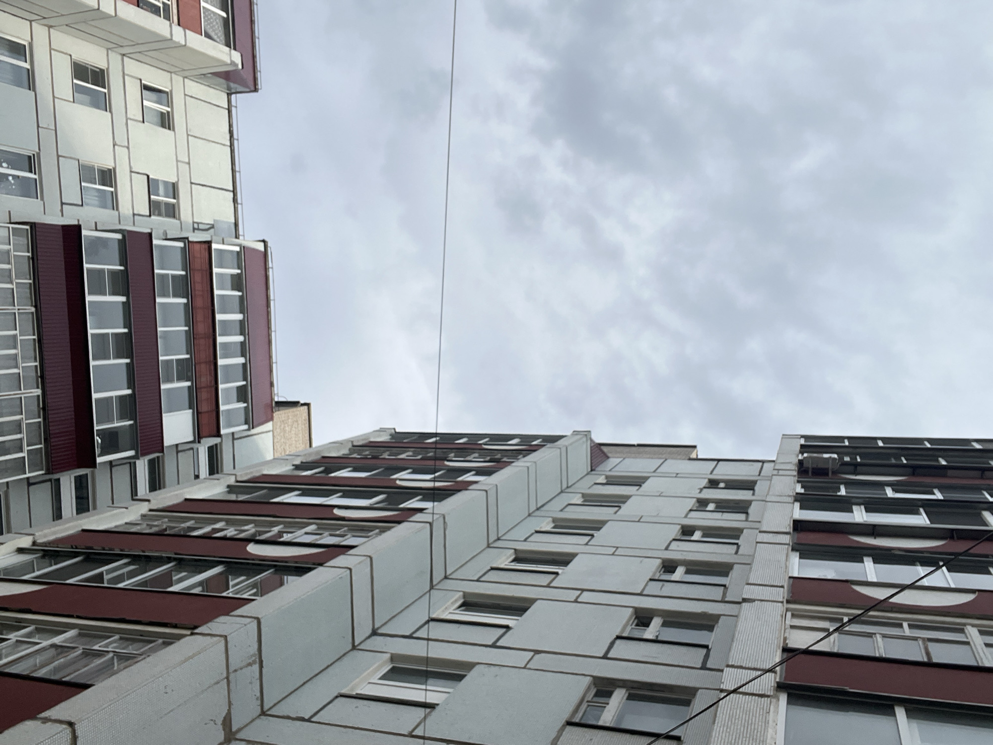 The image depicts an upward-angled view of apartment buildings in Chaikovsky, Russia, under a cloudy sky. The buildings are multi-story structures with a consistent architectural style characterized by rows of windows and balconies. One building in the foreground on the left has a light-colored facade with prominent vertical and horizontal lines, and a band of dark red material along its upper sections. The building on the right is lighter in color and features more traditional window placements. The sky is overcast with diffused light, and a thin wire runs horizontally across the frame, adding a linear element to the composition. No people or active events are visible in the scene.