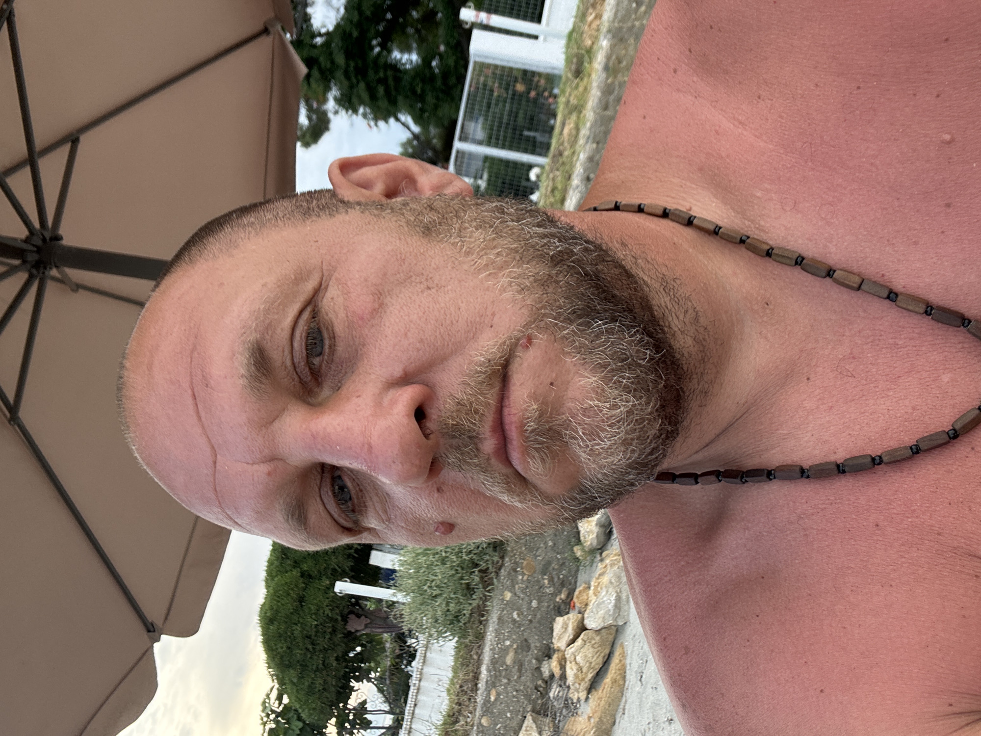 This is a close-up, vertical photograph of a man's face and upper chest, taken outdoors. The man appears to be of Caucasian ethnicity, with short, buzzed brown hair and a beard. He is wearing a wooden beaded necklace. His skin is flushed, suggesting he may have a sunburn. The background includes a portion of a beige umbrella, green foliage, and what appears to be a white fence or railing. The overall lighting suggests it might be late afternoon or early evening. The setting seems to be a casual outdoor area, possibly near a beach or garden, given the presence of stones and vegetation. There is no visible text in the image.