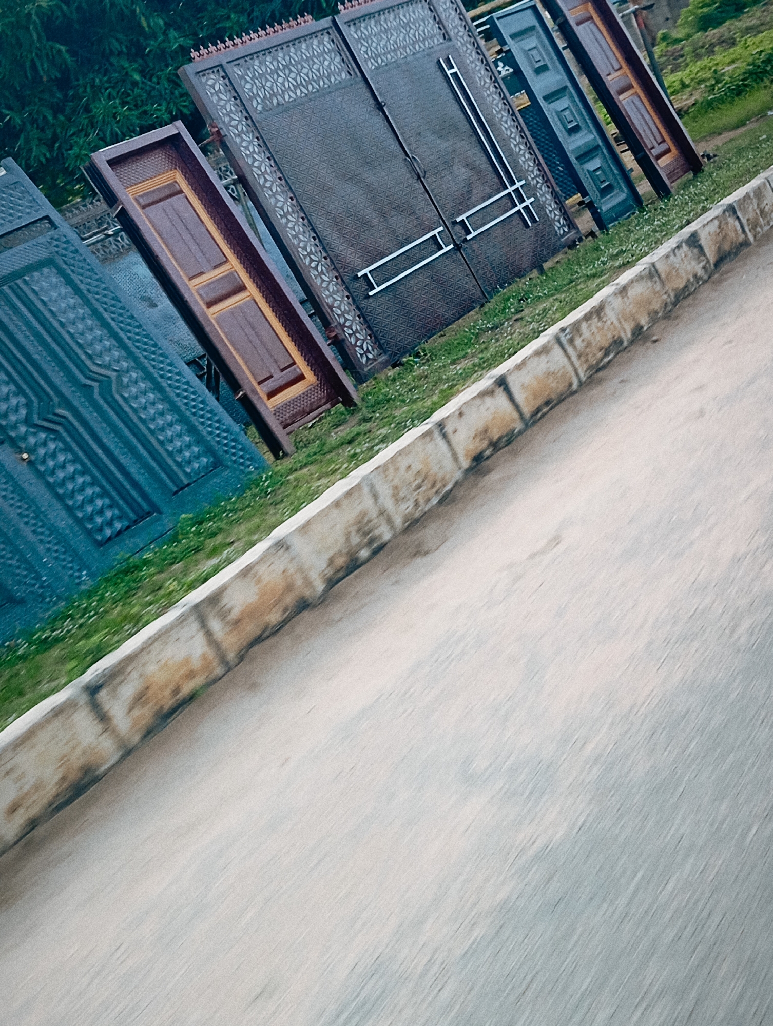 The image depicts a collection of large, decorative gates displayed outdoors, likely for sale. The gates are of various designs and colors, including dark metal with intricate patterns and a dark brown wooden gate. They are positioned at an angle along a grassy area next to a paved surface and a curb made of stone blocks. In the background, there are green trees and foliage. The scene appears to be a roadside display, possibly in Jalingo, Nigeria, where such gates are commonly sold for security and aesthetic purposes for homes and businesses. The lighting suggests it is daytime.