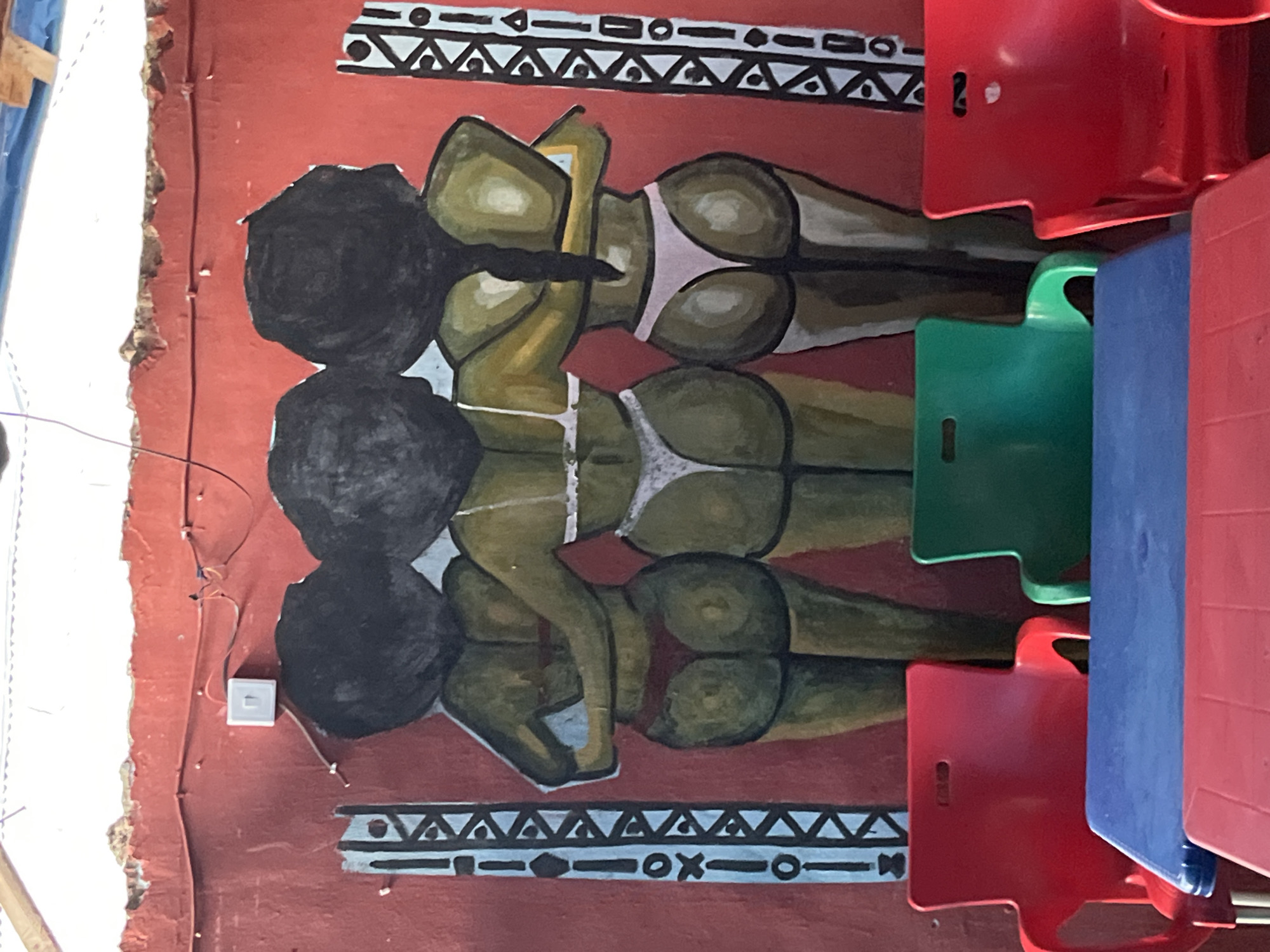 A mural of three women, depicted from behind and wearing bikinis, adorns a red wall in Unknown, Unknown. The women have dark hair and their bodies are painted in shades of green and brown. Decorative blue and white tribal patterns frame the mural on either side. In the foreground, two red plastic chairs and one green plastic chair are visible, along with a blue and red plastic table. The wall above the mural shows signs of wear, with peeling paint and exposed plaster. Electrical wires and a white light switch are mounted on the upper portion of the red wall.