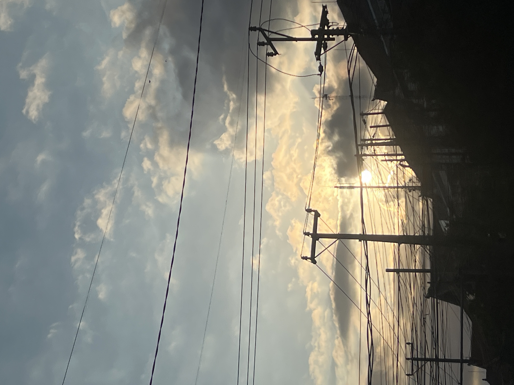 The image depicts a densely populated urban environment in Denpasar, Indonesia, characterized by a profusion of overhead power lines and utility poles silhouetted against a dramatic sky. The sun is setting or rising, casting a warm, golden glow through scattered clouds, some of which are dark and brooding, suggesting an approaching storm or the remnants of one. The foreground is dominated by the intricate network of wires, creating a sense of urban complexity and possibly neglect. Structures, likely buildings or houses, are visible in silhouette at the lower portion of the image, their roofs and architectural details obscured by the low light. No people are clearly discernible in the frame, but the sheer density of the power infrastructure implies a significant human presence and activity within this area of Denpasar. The scene captures a moment of transition in the day, with the interplay of light and shadow highlighting the industrial and residential fabric of the city.