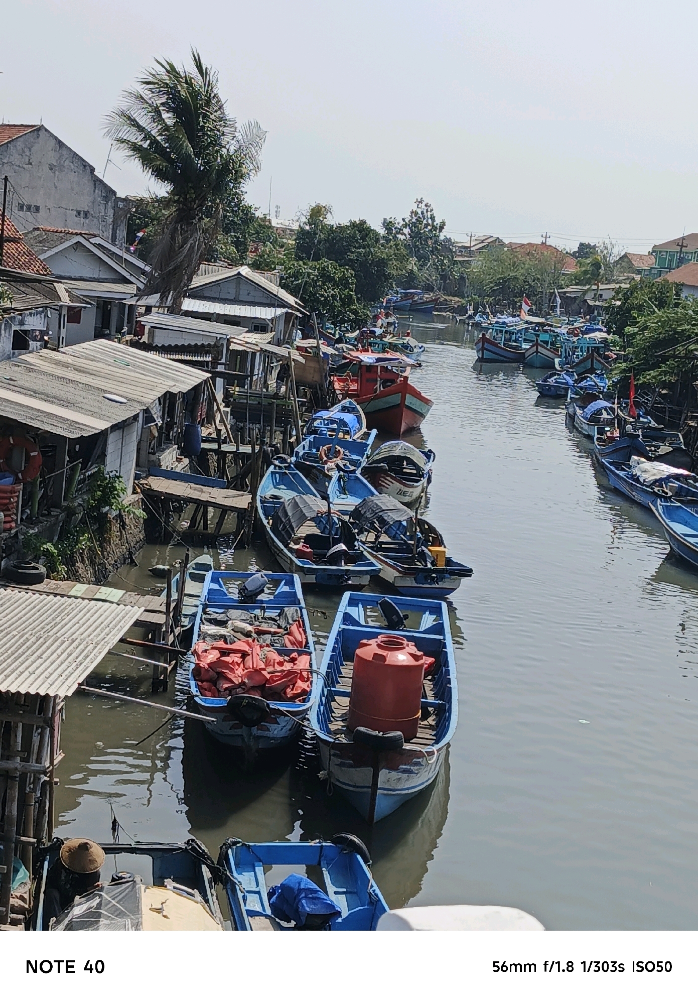 In the city of Kelurahan Cilacap, Kecamatan Cilacap, Indonesia, a waterway is filled with numerous small boats. The boats are predominantly blue and are moored in a tight formation along a canal, suggesting a busy harbor or dock area. Some of the boats are loaded with orange life vests, indicating preparation for fishing or transport. The surrounding environment features a mix of low-rise buildings and dense vegetation, with a prominent coconut palm tree visible on the left side of the frame. The water appears calm, reflecting the overcast sky, which contributes to a muted color palette of the scene. The composition suggests an elevated viewpoint, looking down onto the canal and boats.