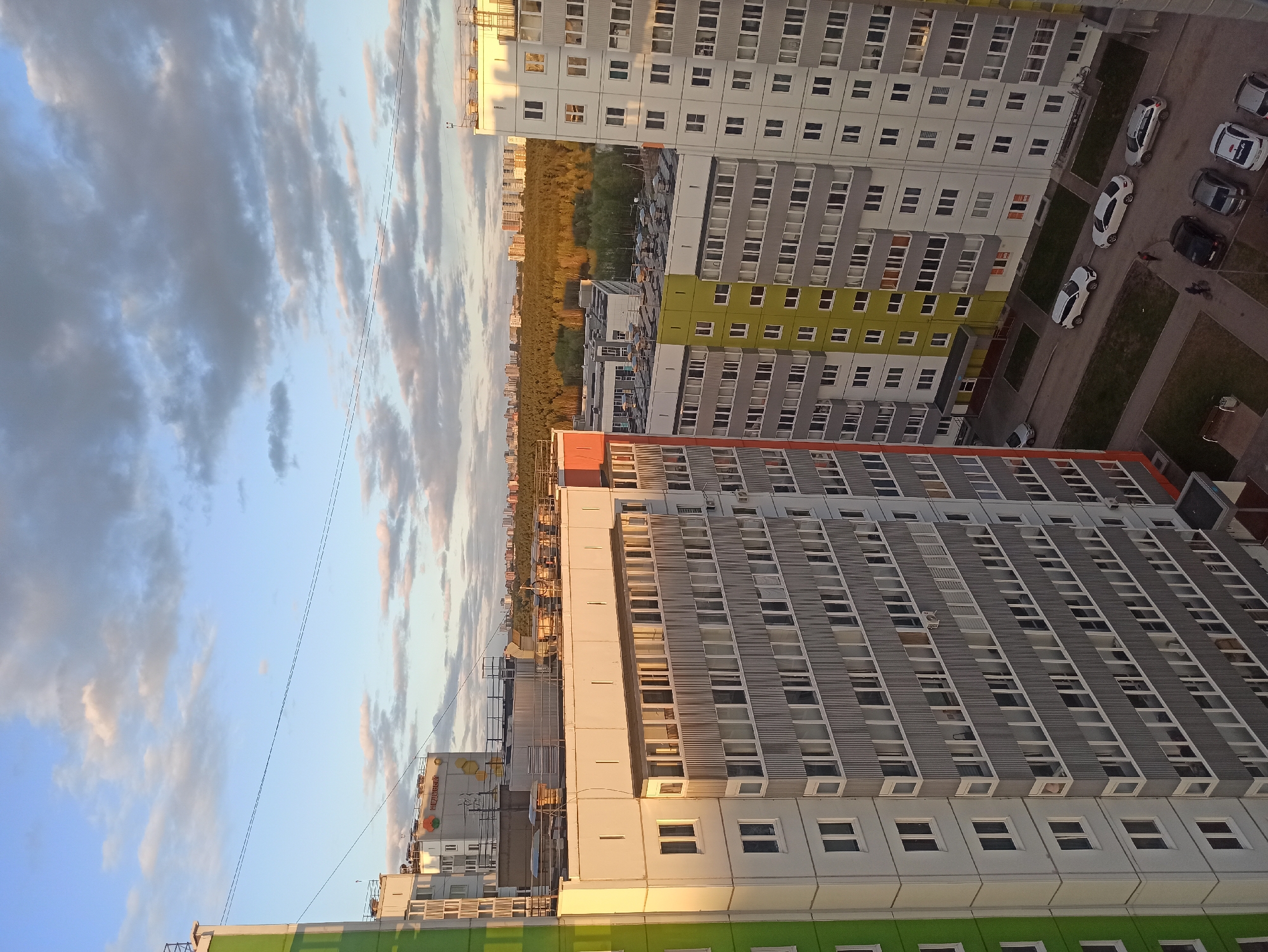 The image shows a high-angle view of apartment buildings in Кондратово, Russia, taken during the late afternoon. The buildings are primarily white, gray, and green, with multiple windows. A row of parked cars is visible between the buildings. In the distance, a line of trees marks the horizon. The sky is partially cloudy with blue patches. Power lines are visible in the sky. The sunlight casts shadows on the buildings, suggesting the photo was taken near sunset.