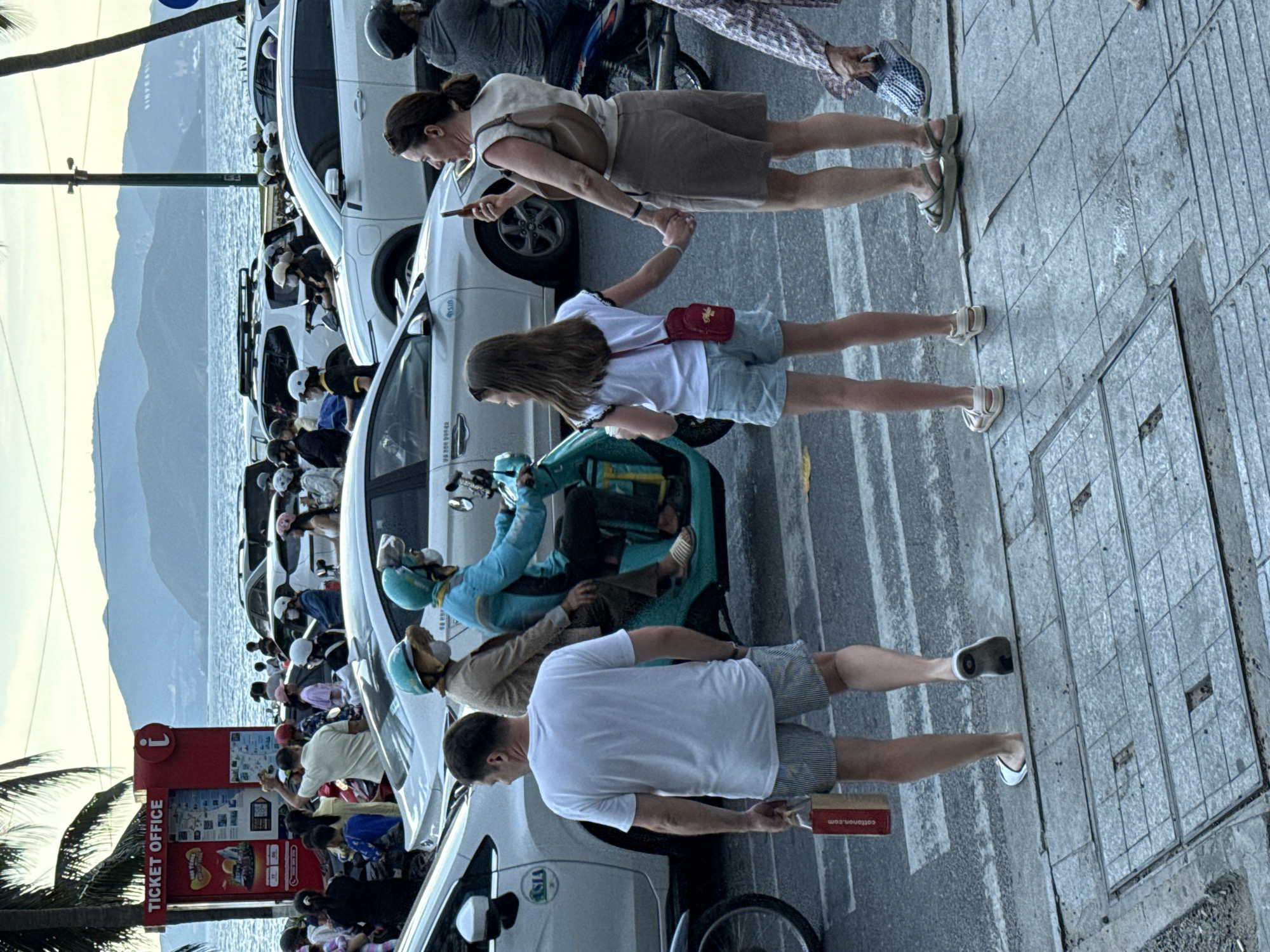 In Nha Trang Ward, Vietnam, a street scene unfolds with a variety of vehicles and pedestrians. A man in a white t-shirt and striped shorts walks across a crosswalk, holding a red paper bag. To his right, a woman and a young girl, holding hands, are also crossing the street. The woman is holding a smartphone and appears to be taking a photo. Behind them, a scooter with two passengers, both wearing helmets and masks, is stopped in traffic. The scooter's passenger wears a teal jacket and is looking forward, while the driver wears a brown jacket and hat, also looking forward. In the background, several cars, including a white sedan with "ASIA" visible on its side, are parked or moving along the street. A red ticket office booth stands to the left, with palm trees and a body of water with distant mountains in the backdrop. The sky is overcast, suggesting late afternoon or early evening. The overall scene depicts a busy urban environment with a mix of local residents and possibly tourists navigating the street.