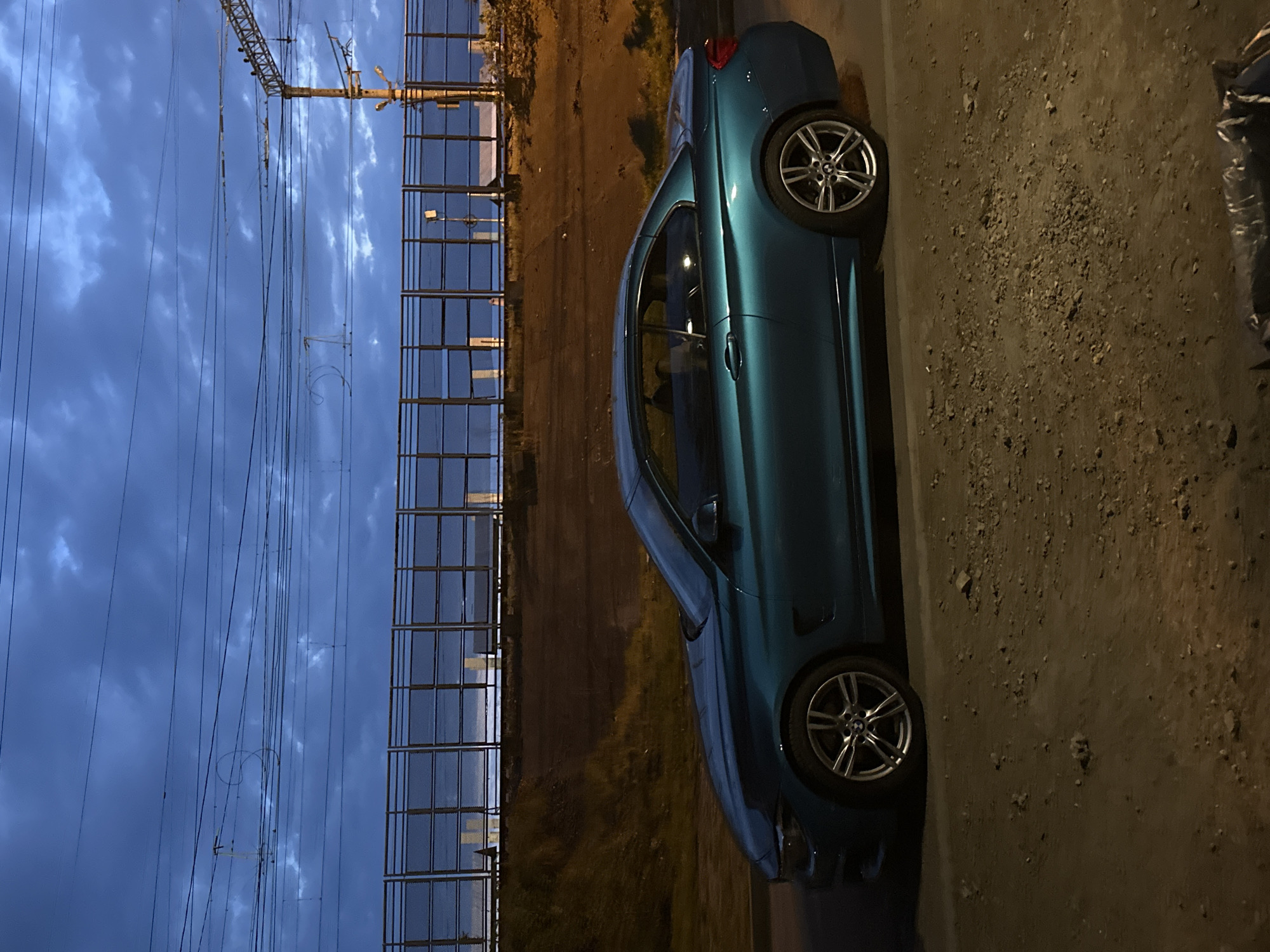 A teal or deep blue BMW car, likely a coupe or sedan, is parked on a gravelly dirt shoulder during twilight or early evening. The car is oriented with its front towards the left and its rear towards the right, showcasing its side profile. Both front and rear wheels are visible, featuring silver multi-spoke rims with the distinct BMW logo on the hub caps.
To the left of the car, a tall, multi-sectioned metal fence or barrier, possibly a sound or security fence, extends horizontally across the frame. Its reflective surface catches the light. Beyond this fence, against a deep blue, cloudy sky, an extensive network of overhead power lines and large power pylons can be seen. The sky is characterized by scattered, elongated clouds.
The ground where the car rests is rugged, composed of loose gravel and dirt, and appears to be illuminated by artificial light, contrasting with the fading natural light in the sky. The scene depicts an outdoor setting, likely adjacent to a road or railway infrastructure, given the prominent power lines and barrier. No individuals are present, and the car is stationary.