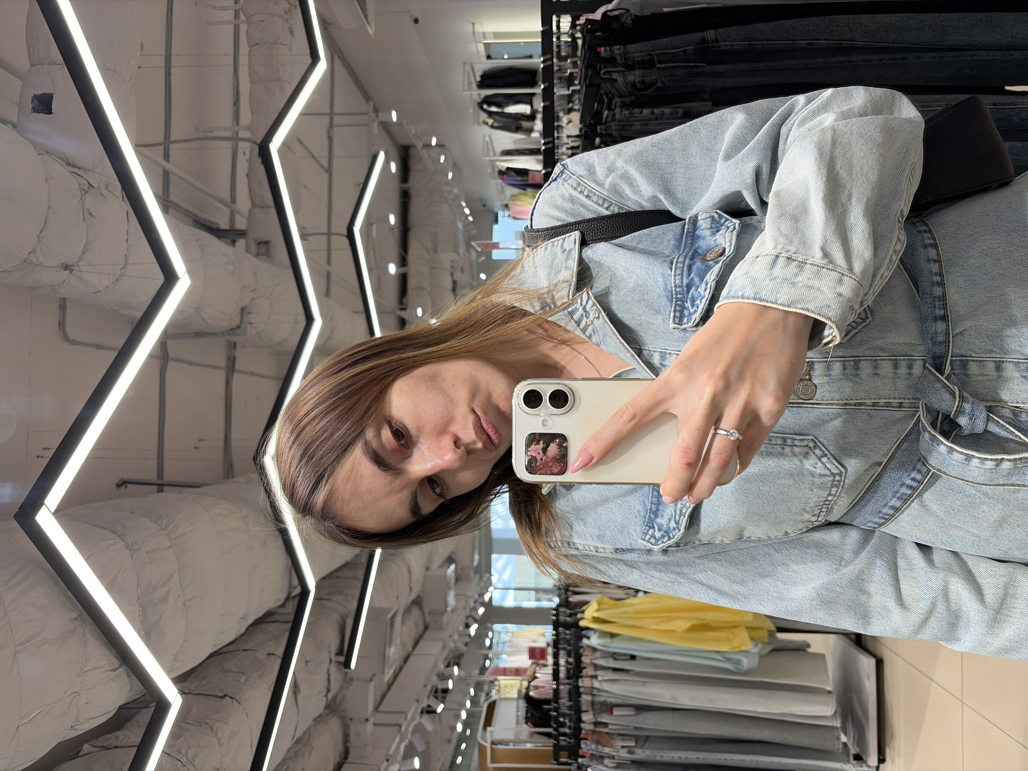 A woman is taking a selfie in a store, likely located in Izhevsk, Russia, as per the query request. The selfie shows the woman holding a white smartphone. She is wearing a light blue denim jacket. A black purse strap is visible over her shoulder. The background is a retail setting, with clothing racks stocked with merchandise. Overhead, the ceiling has exposed ductwork with geometric light fixtures. Garment racks are stocked with clothing, including a section of yellow shirts/sweaters and a section of grey pants.