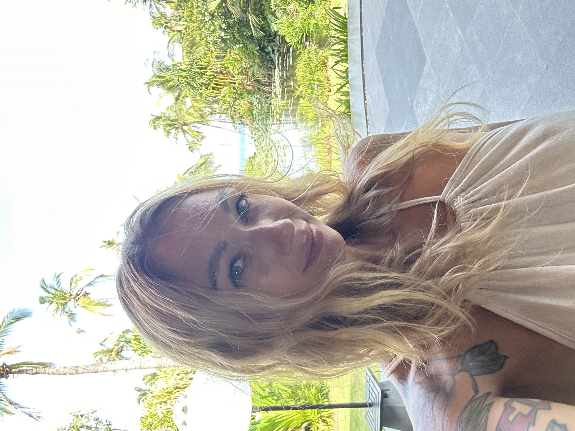 A blonde woman with tattoos on her left arm and shoulder is taking a selfie outdoors. She is wearing a beige halter dress. The background shows a pond with lily pads, lush green vegetation, palm trees, and a hint of the ocean in the distance. The location is Phu Thuy Ward, Vietnam.