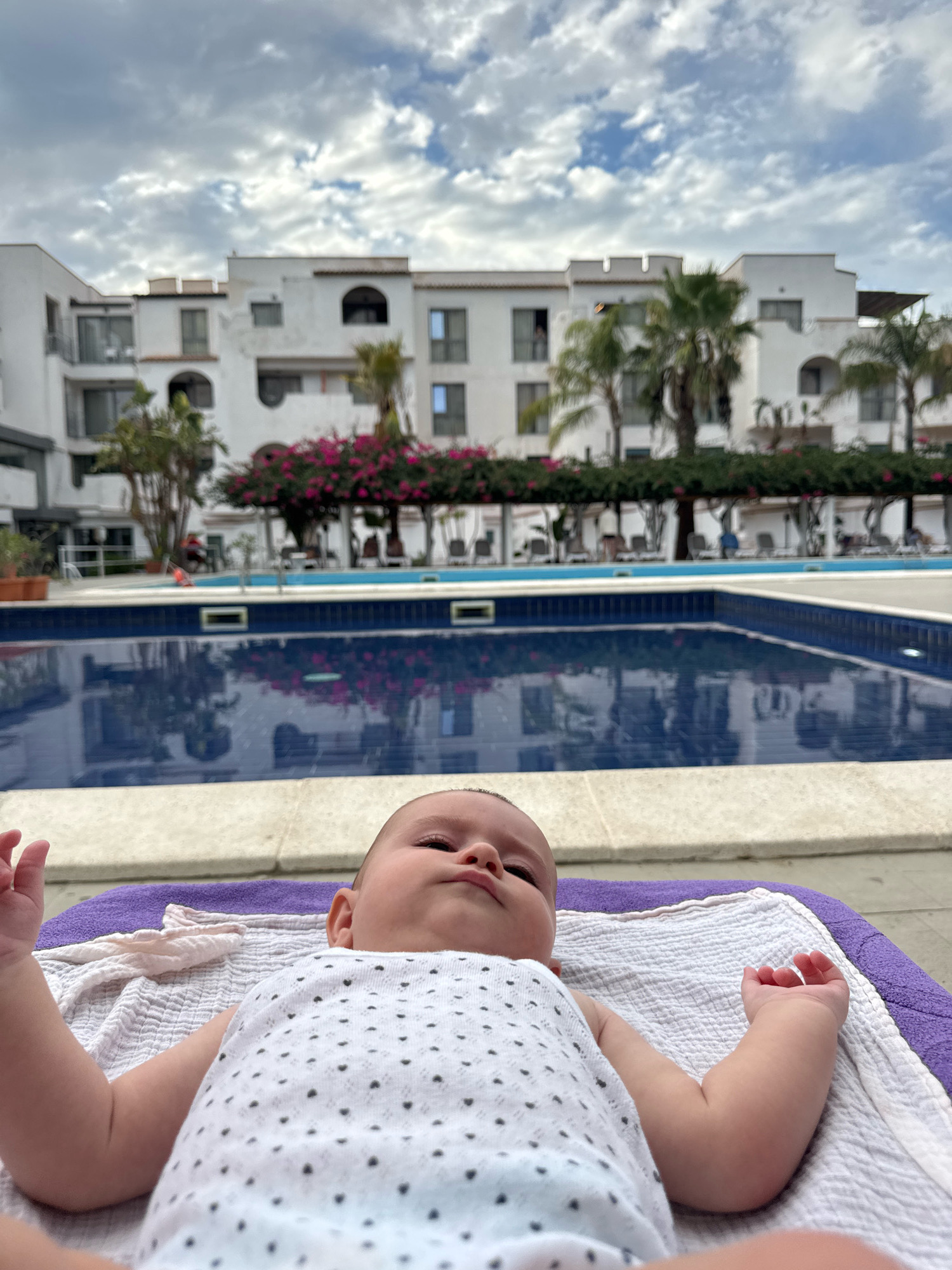A close-up, low-angle shot shows a baby lying on a purple towel and a white textured blanket. The baby is wearing a white onesie with black polka dots. The baby's head is tilted back, and they are looking up towards the sky.
In the background, a swimming pool with blue water reflects the surroundings. Beyond the pool, a white building with multiple floors, balconies, and windows is visible. Palm trees and flowering plants line the area around the pool and in front of the building. The sky above is partly cloudy with white and gray clouds. The overall scene appears to be a resort or hotel environment, possibly located in Isola delle Femmine, Italy, given the architecture and vegetation.