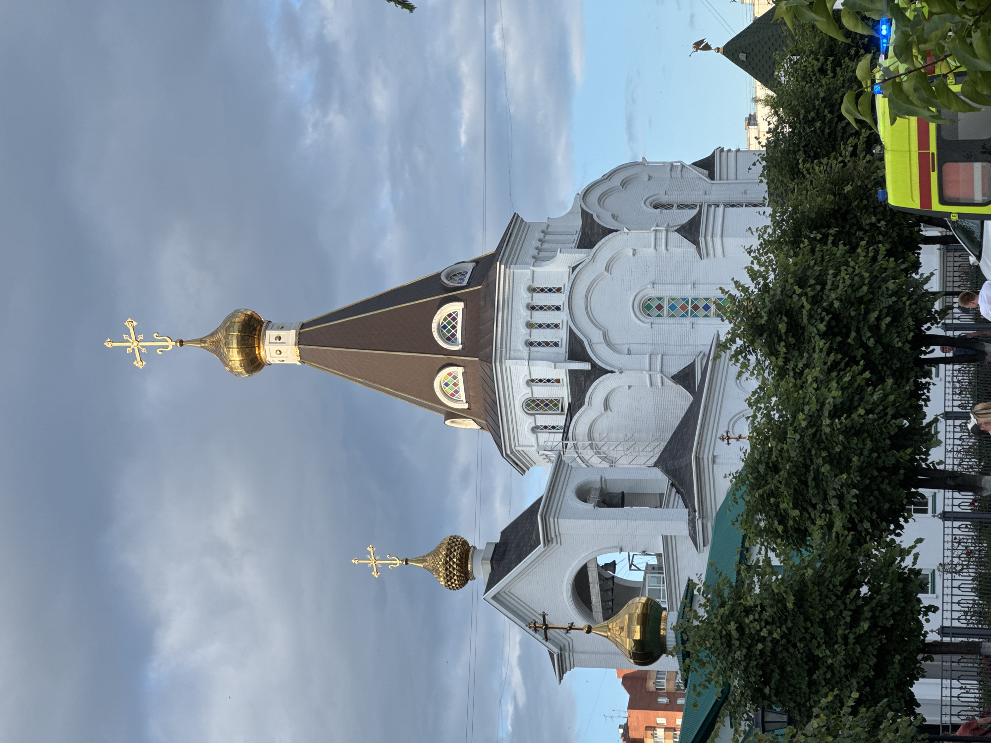 A tall, white, Orthodox church with multiple golden domes and crosses stands against a cloudy sky. The main dome is conical and dark brown, topped with a golden cross. To the left, a smaller golden dome sits atop a white structure with an arched opening containing bells. The church has arched windows with stained glass. In the foreground, lush green trees partially obscure the view, and a bright yellow ambulance with flashing blue lights is parked behind a black metal fence. Several people are visible in the distance, near the fence. The overall scene suggests an urban setting, possibly in Russia or another Eastern European country, given the architecture.