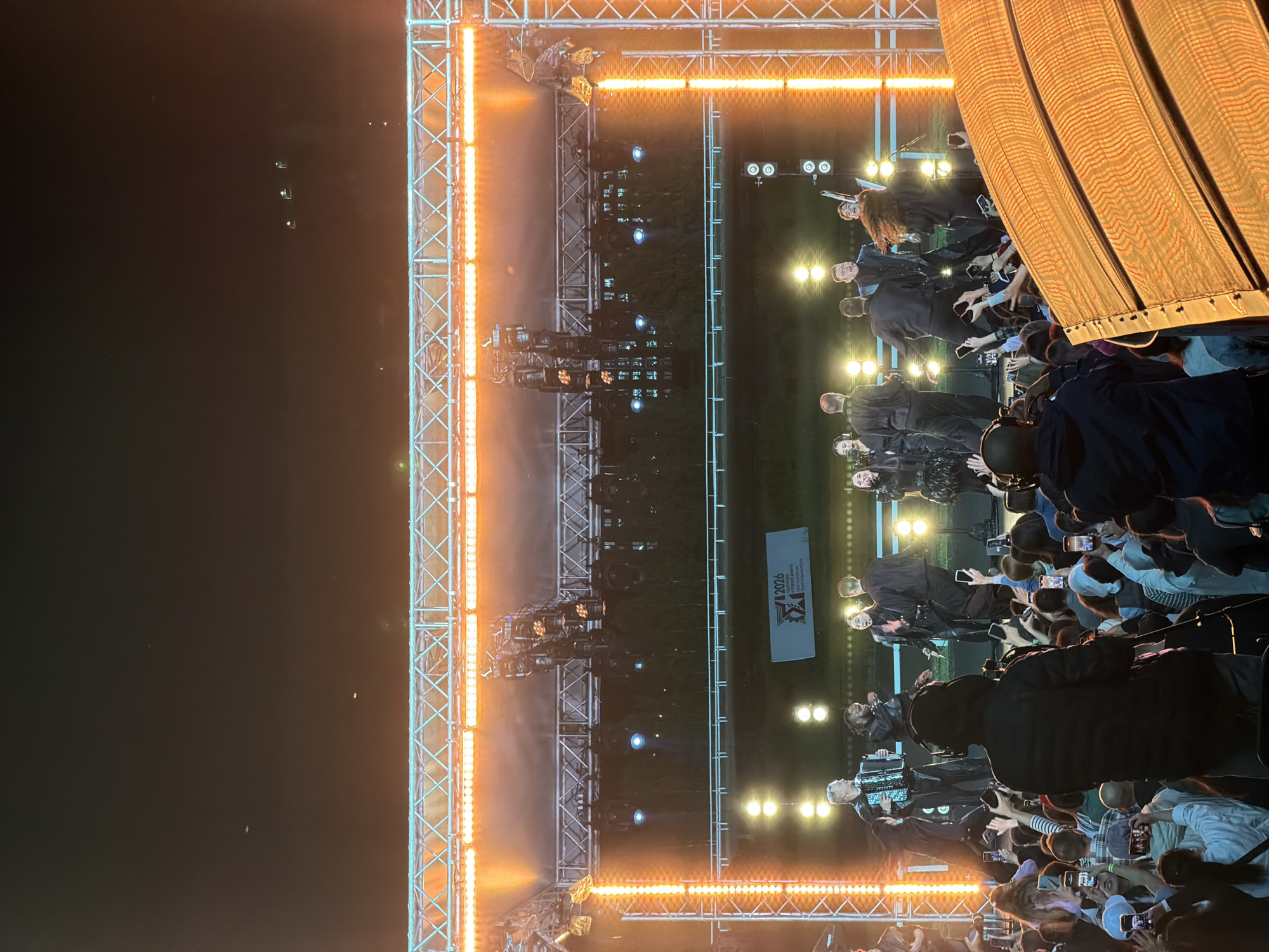 An outdoor concert is underway at night, with a stage lit by orange and white lights. A group of performers is on stage, including a man playing an accordion and several singers. A crowd of people is gathered in front of the stage, many of them holding up their phones to record the performance. The concert appears to be taking place in a park or open field, with trees and a building visible in the background.