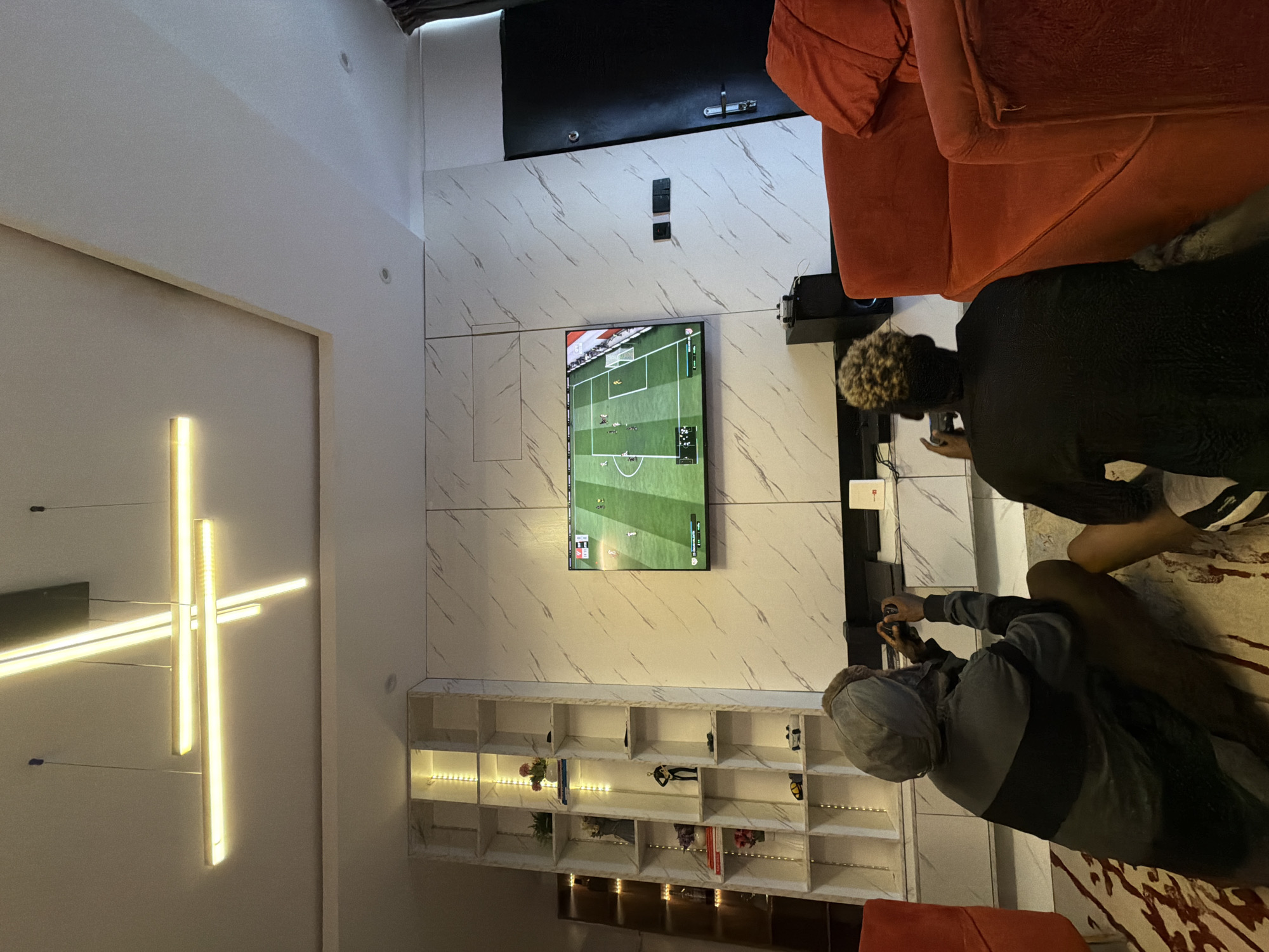 Two people are playing a video game on a television in what appears to be a living room. The wall behind the TV is decorated with marble-patterned paneling. Below the TV, a shelf unit displays various decorative items. One person is wearing a grey hoodie, while the other is wearing a black shirt. The room has an orange couch and a modern lighting fixture. The location is in Igboefon, Nigeria.