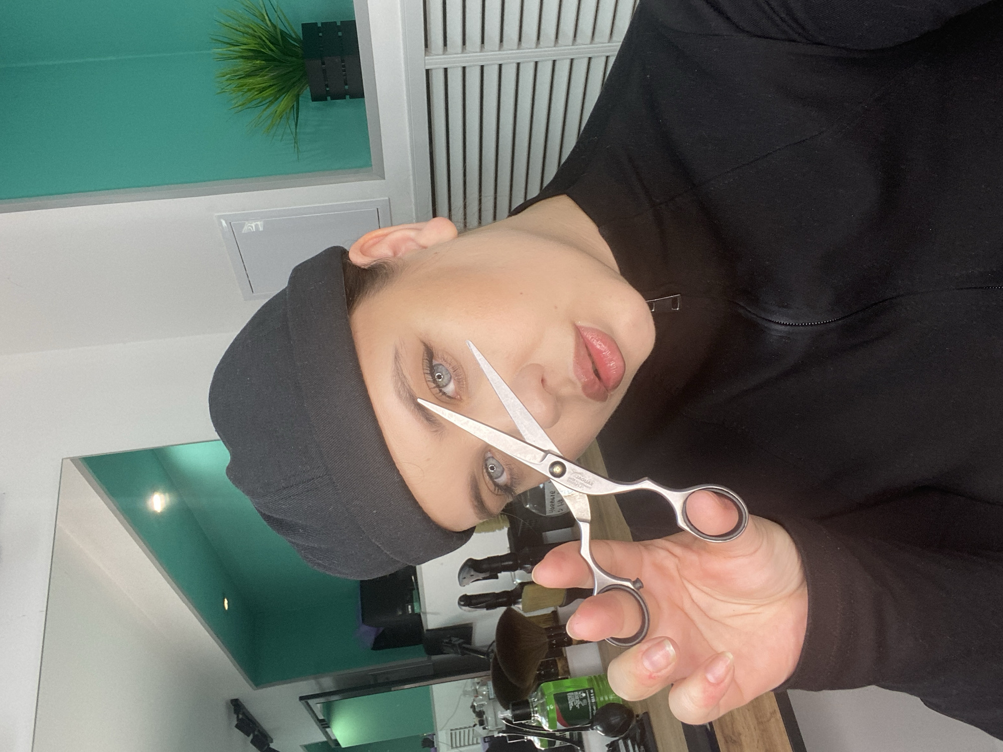 A person wearing a black beanie and black clothing is holding a pair of scissors in their right hand. The person's face is visible, and they have light eyes. The scissors are silver and appear to be professional hairdressing shears.
The background includes a turquoise wall, a window with blinds, and a potted plant near the top of the frame. Below the person's hand, there are some styling tools and products visible, partially obscured by the person and the scissors.
There is no evidence to support that this image was taken in Chernushka, Russia.