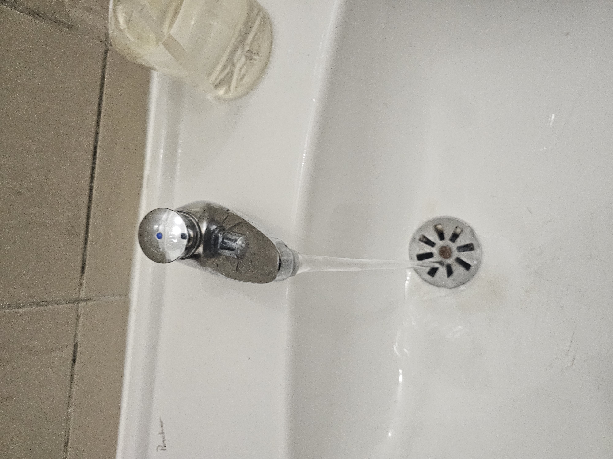 A top-down shot shows a sink with a faucet running water down the drain. The sink appears to be white and made of porcelain. The faucet is made of metal and has a blue and silver handle. Water is running from the faucet into the sink drain, which has a flower-like design. A soap dispenser is partially visible in the upper left corner of the frame. The sink is situated next to tiled walls. The word "Rocher" is faintly visible near the sink's edge, suggesting the brand of the sink. The scene is likely in a bathroom or washroom environment. Due to the lack of additional information, the location in Paris, France, cannot be determined.