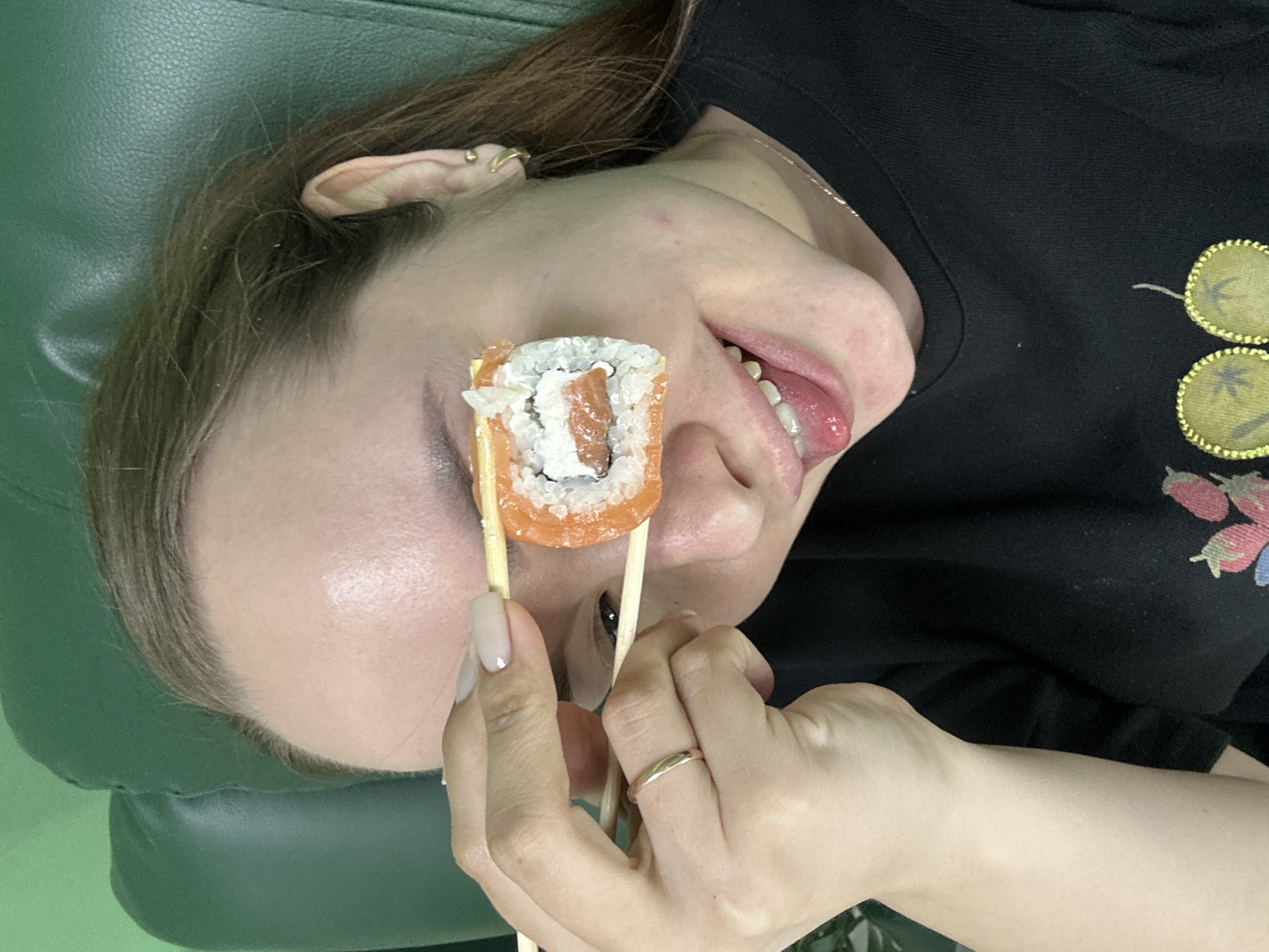 A woman is lying on what appears to be a green upholstered surface, possibly a couch or chair. She is holding a sushi roll with chopsticks in front of her eye. The sushi roll is cylindrical and consists of an orange outer layer, likely salmon, with white rice and a dark ingredient inside. The woman is wearing a black shirt with a floral pattern. She has a gold ring on one of her fingers and long, light-colored nails. She is wearing jewelry in her ear. There is no visible information to confirm the location of the scene to be Chaikovsky, Russia.