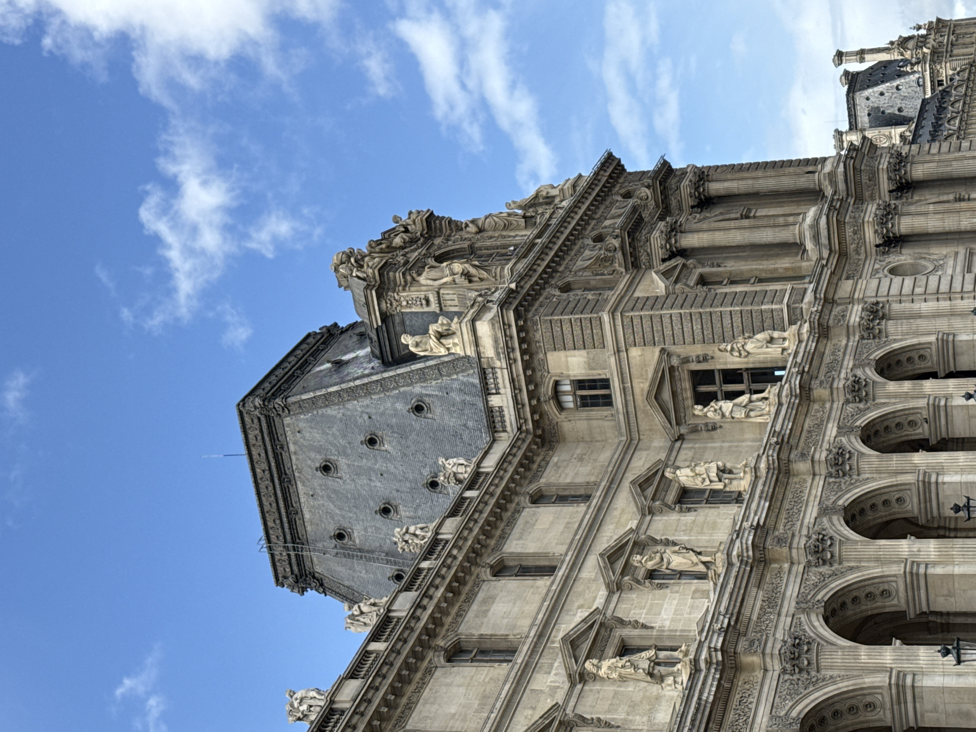 The image captures a close-up, angled view of a section of the Louvre Palace in Paris, France. The architecture is heavily ornamented with classical features including columns, arched windows, and numerous sculptures of human figures adorning the facade. The building material appears to be light-colored stone or concrete, weathered and aged.
The roof is covered with dark, patterned tiles or slates, punctuated by dormer windows and smaller decorative elements. A clear blue sky with scattered white clouds provides the backdrop. The angle of the shot emphasizes the height and grandeur of the structure, as well as the intricate details of its construction and decoration.