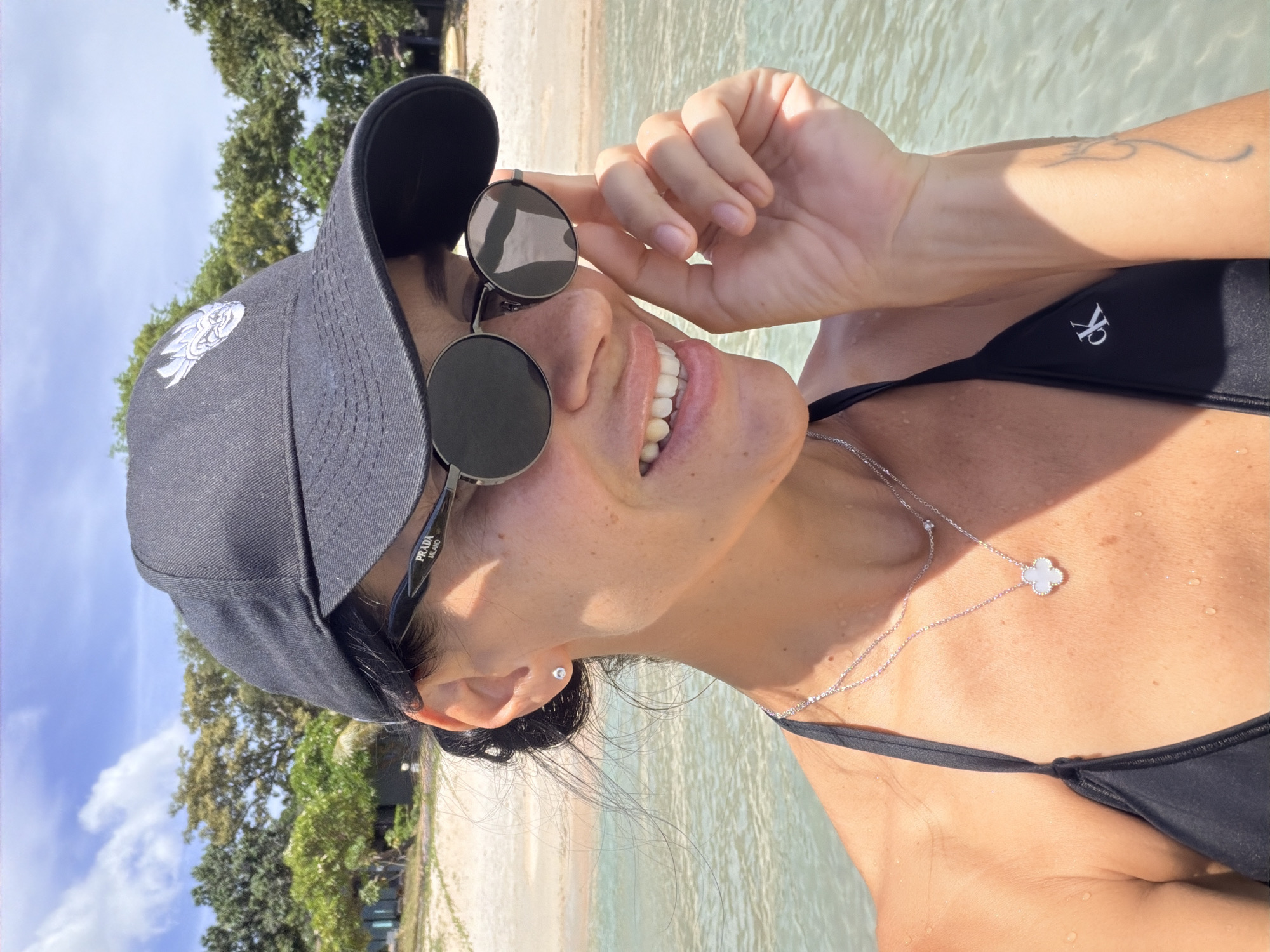 A woman is taking a selfie in the ocean in Ko Samui District, Thailand. She is wearing a black baseball cap, black sunglasses, and a black bikini top. She is also wearing a silver necklace with a clover-shaped pendant. Her left hand is touching her sunglasses. The background shows a sandy beach with palm trees and clear blue water.