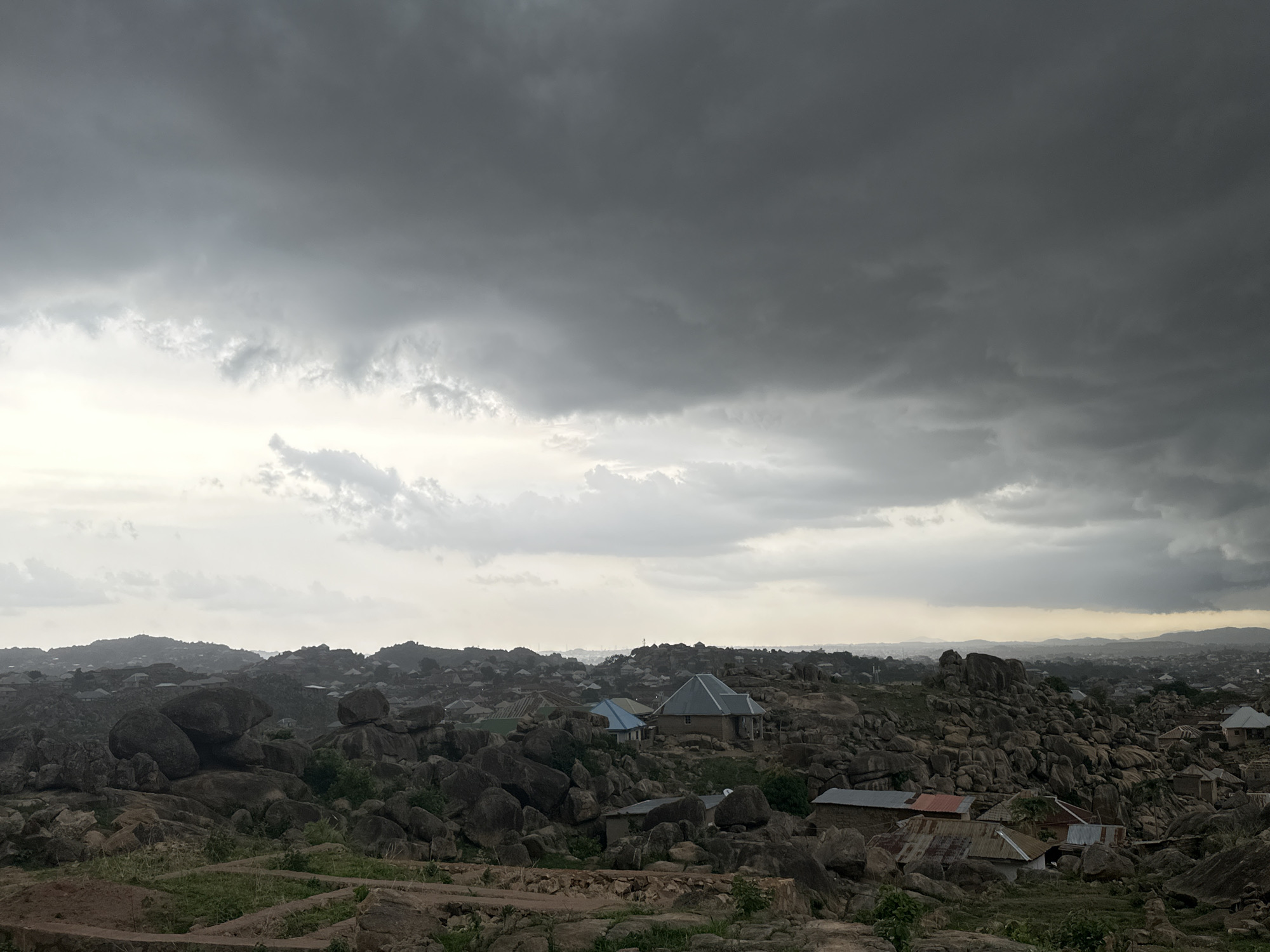 The image shows a landscape in Jos, Nigeria, under a heavily overcast sky. The foreground and midground are dominated by large, rounded rock formations interspersed with sparse vegetation and scattered buildings. The buildings appear to be simple, single-story structures with corrugated metal roofs, some showing signs of wear. In the distance, a hazy horizon reveals more buildings and terrain, suggesting a densely populated area. The overall tone of the scene is subdued due to the grey, cloudy conditions. There are no visible people or animals. The primary subjects are the natural rock formations and the built environment integrated within them.