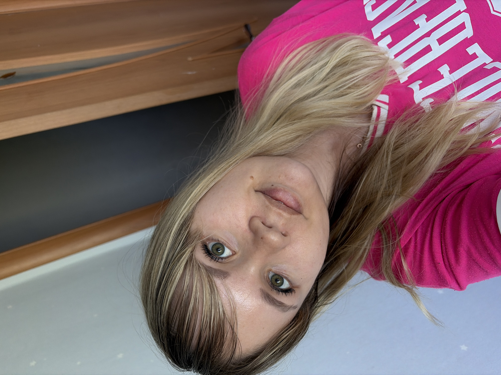 A blonde-haired woman is shown in a selfie, wearing a pink shirt with white lettering. Her eyes are green, and she appears to be indoors. The background includes a wooden structure and a light-colored wall with star patterns. There is no verifiable information in the image that indicates the location is the city of Chaikovsky, Russia.