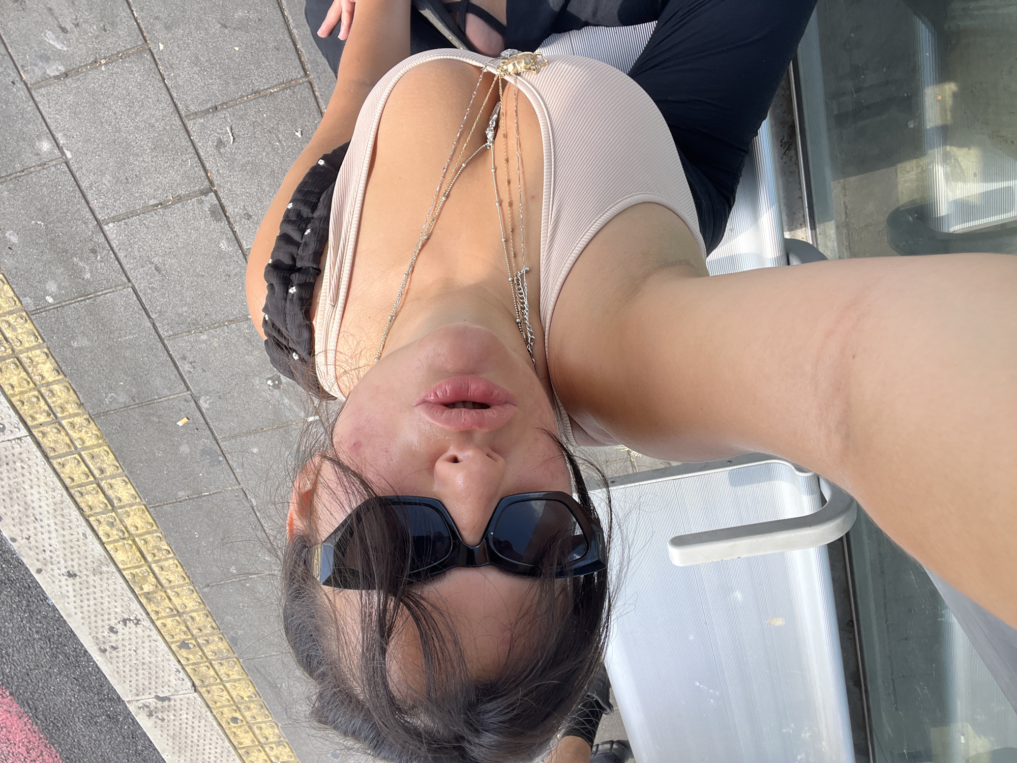 A young woman with dark hair and black sunglasses is taking a selfie outdoors. She is wearing a beige tank top and a black ruffled skirt or top layer. Multiple delicate necklaces are visible on her chest. She is seated on a silver bench. The background shows a tiled sidewalk and a street with yellow tactile paving. The lighting suggests it is daytime. The image appears to be taken in Kiryat Motzkin, Israel, possibly near a bus stop or public seating area.