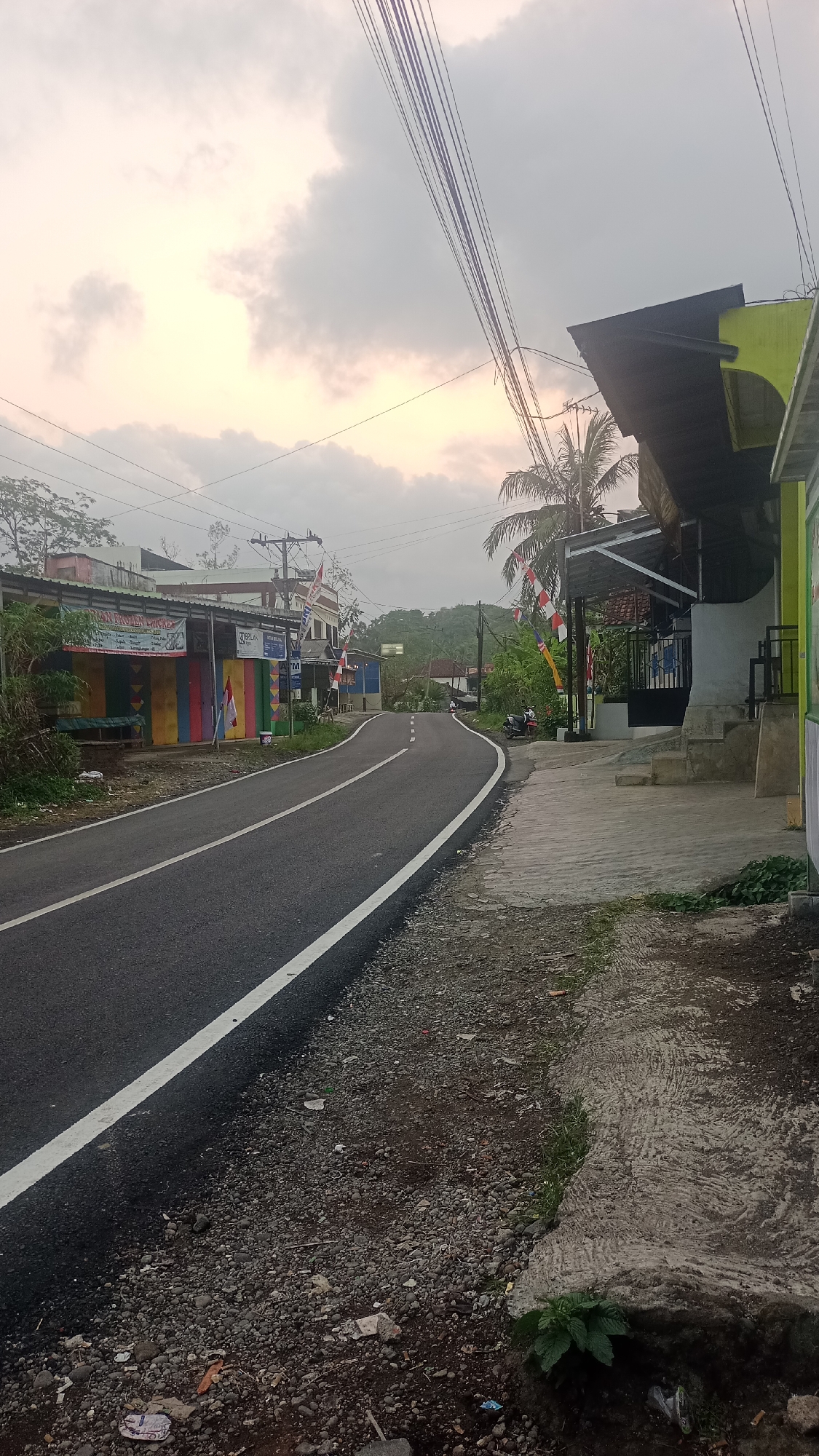 The image depicts a paved road in a village or rural area, likely in Cogreg, Indonesia, under a cloudy sky with hints of sunrise or sunset light. The road curves to the left and is bordered by a gravel shoulder with scattered debris. On the left side of the road, there are several storefronts with colorful facades, some displaying Indonesian flags. A few motorcycles are parked near these establishments. On the right side of the road, a concrete walkway leads up to a building with a dark roof and yellow trim. A palm tree is visible behind this building. In the distance, more buildings and lush green hills are present. The scene is quiet, with no people actively engaged in any discernible actions. The overall impression is of a developing or established community in a tropical environment.