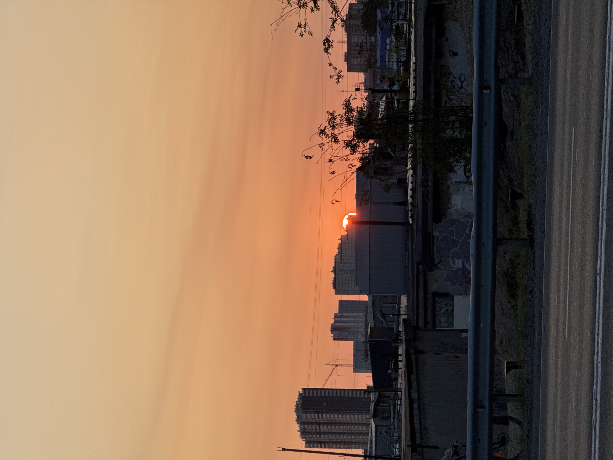 The image depicts a sunset over a city, likely Surgut, Russia, given the presence of industrial buildings and apartment blocks characteristic of the region. The sky is a vibrant gradient of orange and yellow, with the sun partially obscured by a building. Silhouettes of tall apartment buildings and industrial structures dominate the background. In the foreground, a metal guardrail runs along a road. To the left of the guardrail, the wheel and frame of a bicycle are visible. The environment appears somewhat industrial, with low-lying buildings, some exhibiting graffiti, and overgrown vegetation. The overall impression is one of an urban sunset, with a mix of residential and industrial elements.