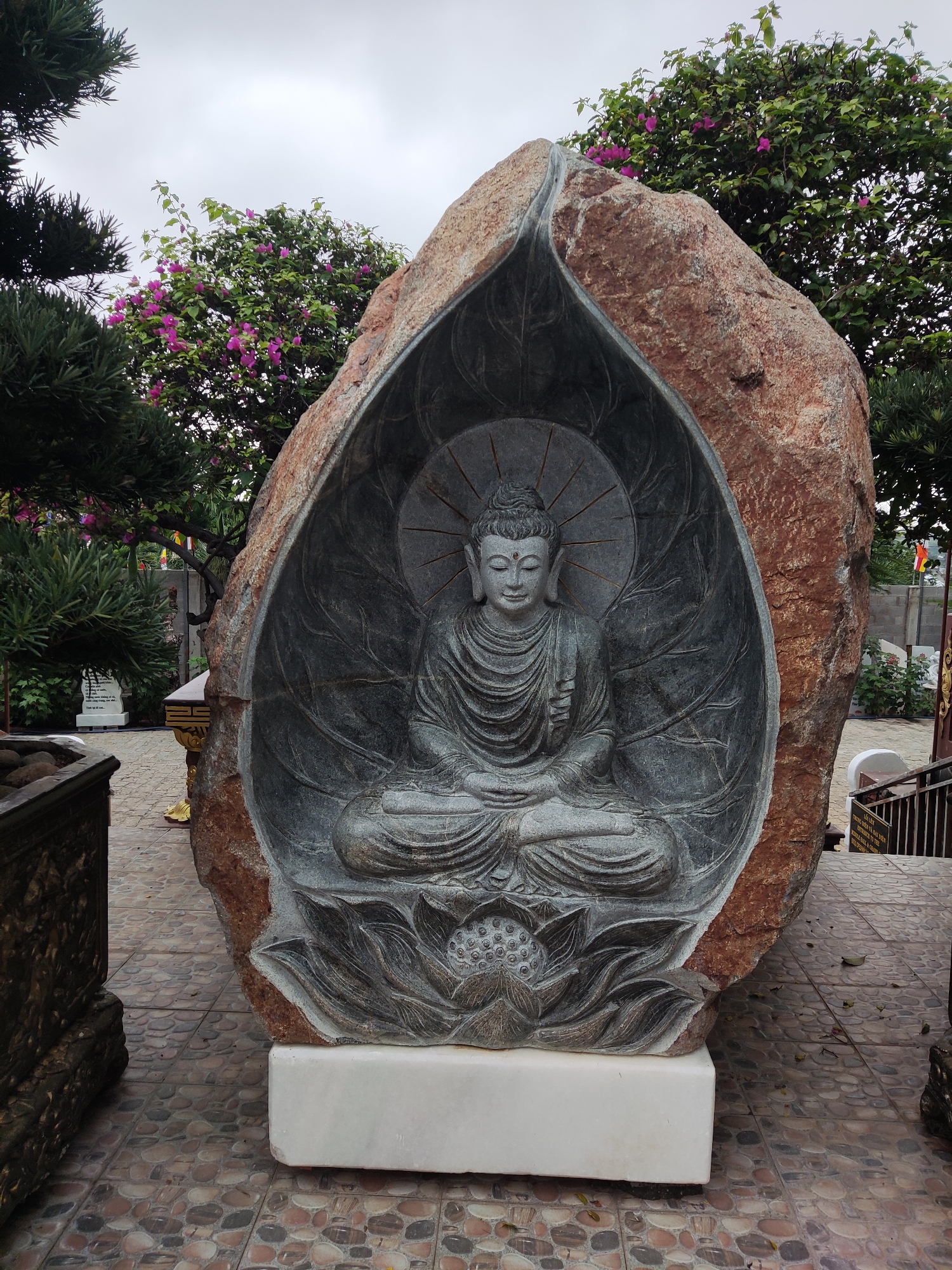 The image displays a serene outdoor scene dominated by a large, carved stone Buddha statue. The grey Buddha, depicted in a meditative lotus position with a peaceful expression, is nestled within a larger, rugged, reddish-brown rock that has been artistically shaped to resemble a protective shell or leaf. Behind the Buddha's head, a radiating halo is carved, and it rests upon a detailed lotus flower base. The entire sculpture is elevated on a white rectangular plinth.
The setting appears to be a tranquil garden or temple grounds. Green foliage frames the sculpture, with coniferous trees on the left and a vibrant bush featuring pinkish-purple flowers (likely bougainvillea) visible against a bright, overcast sky. The ground is paved with an intricate mosaic of small, rounded stones.
In the background on the right, a set of stairs with a dark metal railing can be seen, along with a faintly visible multi-colored Buddhist flag (yellow, white, red, blue, orange stripes). A sign near the stairs displays Vietnamese text, reading, in part, "VÀ ĐÀI SEN TRONG ĐÀI BIỀN NHÂN VỚI THỂ KHOẢN TUYỆT PHÁP VŨ" and "LÒ ĐIỆN PHÁP CHUYỂN PHÁP LUÂN VÔ VÔ VÀ VÔ NGẦM KHẢ XƯNG." The time of day appears to be an overcast day, providing soft, diffused lighting with no harsh shadows. No specific activity is taking place, contributing to the contemplative atmosphere.