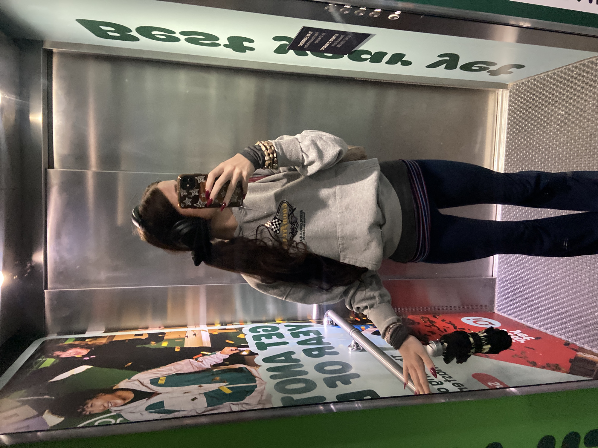 A person is captured in a reflection, likely taking a selfie, within what appears to be an elevator or a space with reflective metallic walls. The image is oriented sideways. The subject, a person with long dark hair, wears a grey hooded sweatshirt with a logo, dark trousers or jeans, and headphones. Their nails are painted red, and they wear multiple beaded bracelets on their right wrist. They are holding a phone with a decorative, patterned case (featuring stars or a monogram-like design).
The setting features polished metallic panels on the walls. The top section and floor are green. On the top section, there is green text on a white background, which appears reversed, reading as "BGZ" and possibly "MAY" or "MAI". A small black sign above this text shows reversed instructions or warnings.
On the floor, there's a large poster featuring two individuals, one prominently wearing a green and white jacket. The poster also contains various reversed text elements, including "GOOD AEV RAY", "GEL VWO", and the number "24". A small, black, folded umbrella or similar object rests on the floor next to the poster. The overall impression is an indoor, modern, and possibly commercial space.