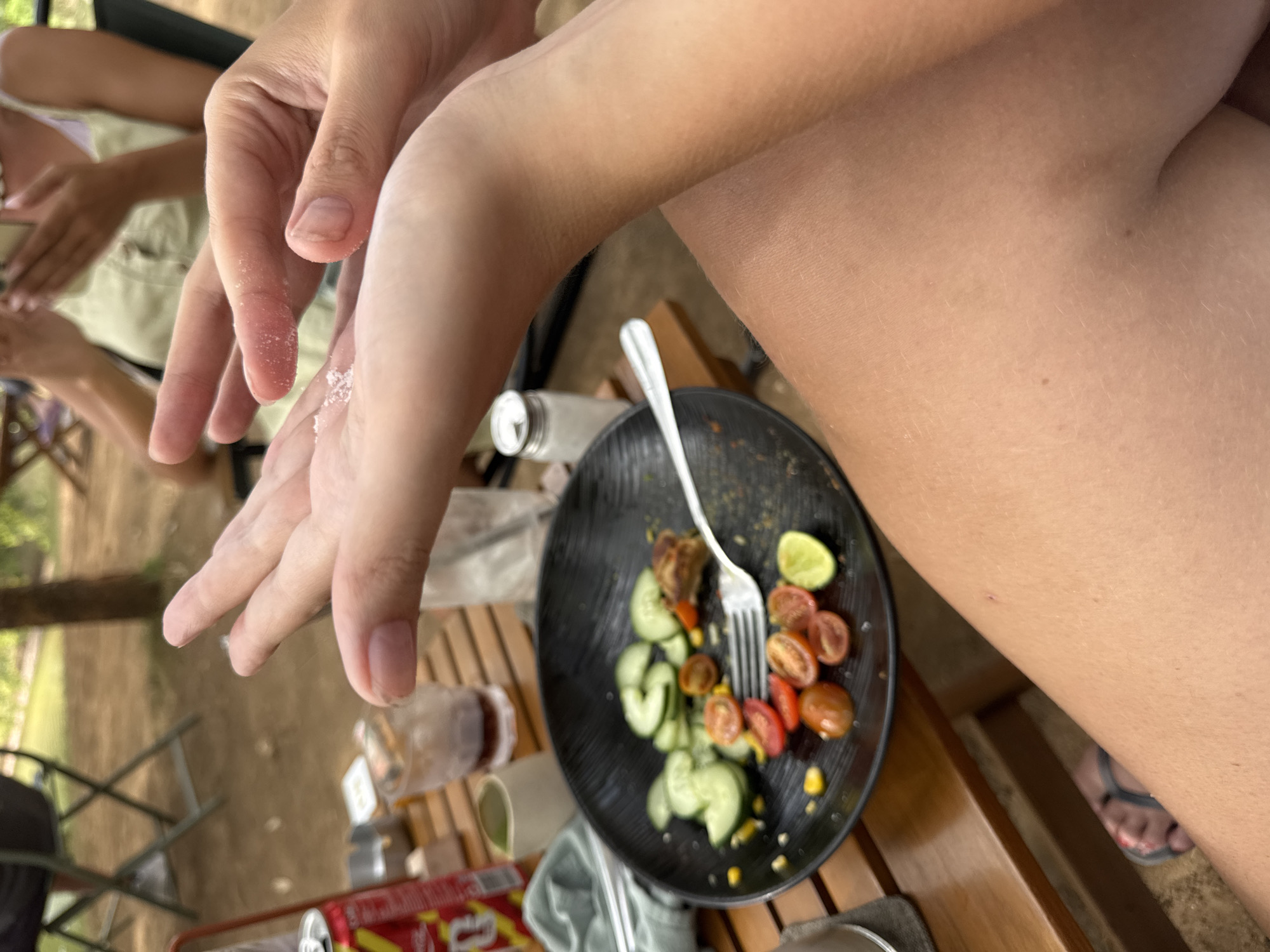 In Kuta Selatan, Indonesia, a close-up, eye-level shot captures hands holding a small pile of white granules, possibly salt or sugar, over a partially eaten salad on a black plate. The salad contains sliced cucumbers, cherry tomatoes, corn kernels, and a piece of meat. A fork rests on the plate, and to the left, a can of Coca-Cola and a napkin are visible. In the background, blurred figures and outdoor seating suggest a casual dining environment. The lighting is natural, indicating an outdoor setting.