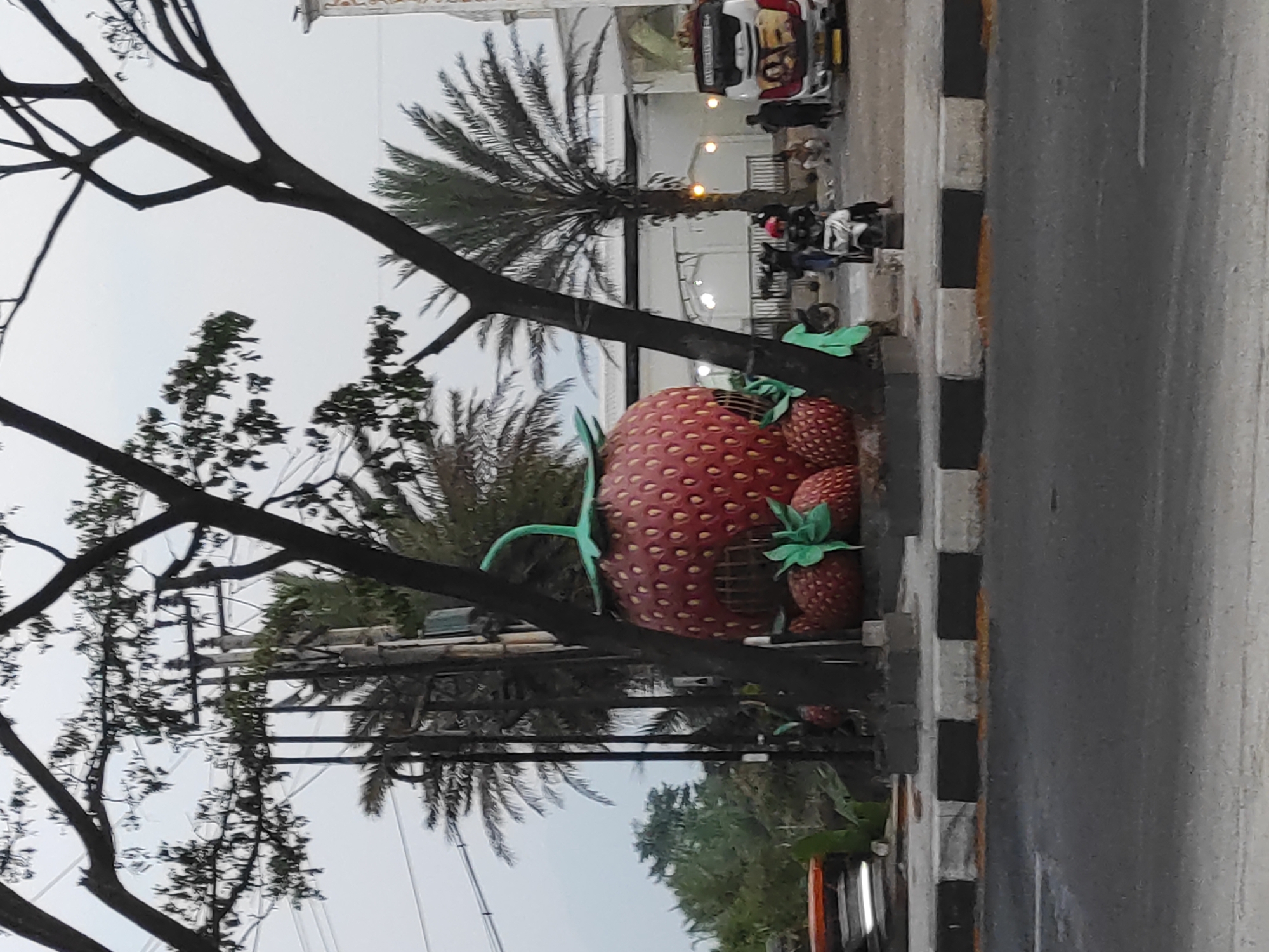 The image captures a roadside scene in Panenjolan, Indonesia. Dominating the foreground is a large, whimsical sculpture of a strawberry with smaller strawberries at its base. The sculpture appears to be a building or an entrance to a venue, with an opening resembling a doorway. Trees with dark branches and sparse leaves frame the sculpture, and in the background, palm trees are visible. Behind the sculpture, a building with white walls and arched openings is situated, and several people are present. One person is on a motorcycle, another is walking, and a few individuals are sitting or standing near a white and red bus with colorful graphics on its rear. The scene is bordered by a road with a black and white striped curb. The sky is overcast.