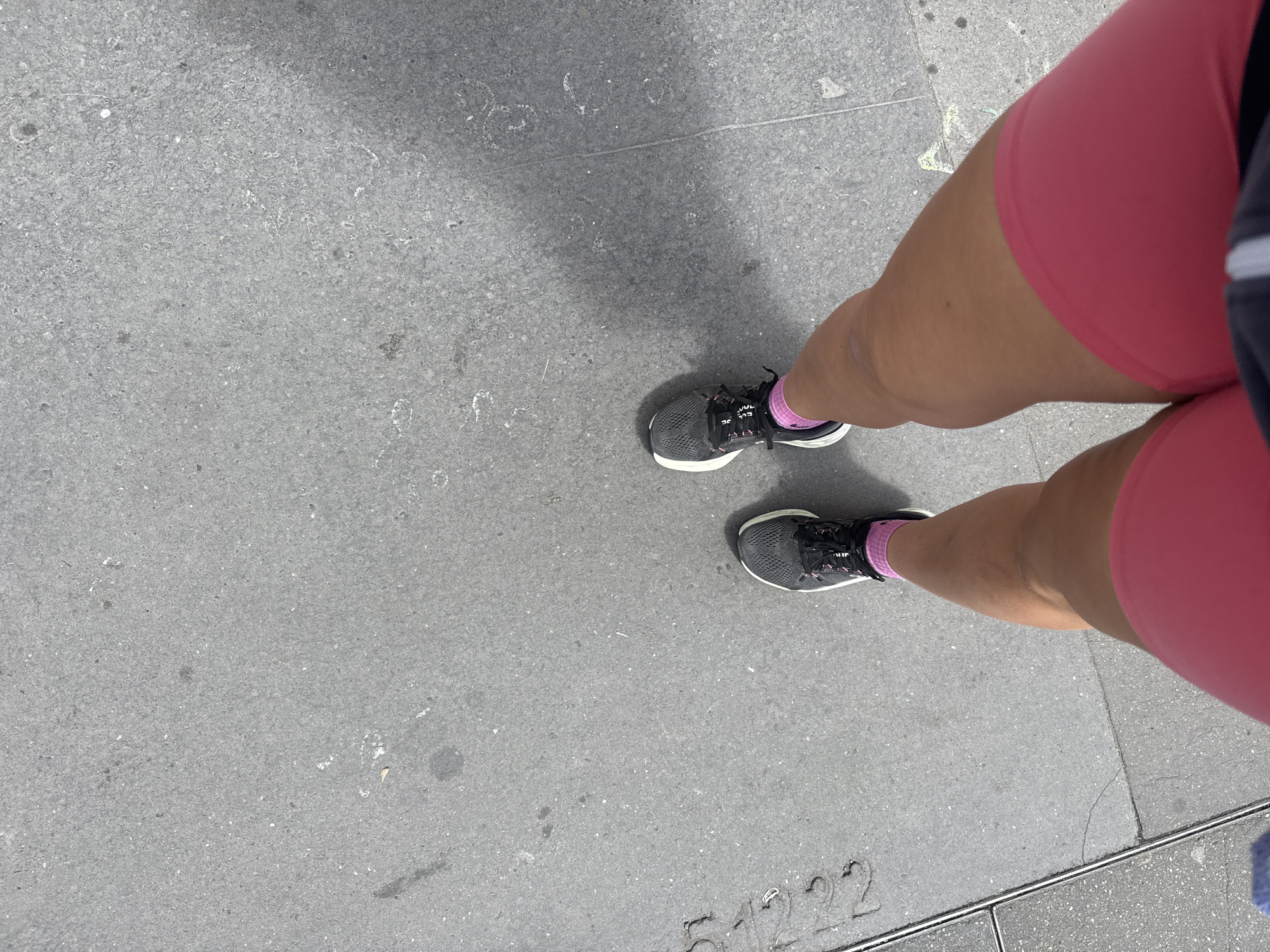An overhead shot shows a person standing on a gray paved surface, likely a sidewalk. The person is wearing pink shorts and gray athletic shoes with pink socks. The surface appears to be concrete or asphalt with some visible markings and slight variations in texture. Lower right shows a recessed metallic bar. The location appears to be an urban setting. The metallic bar is embedded into the sidewalk, and has 51222 printed into the cement next to it. Given this identifier, the location is most likely Paris, France.