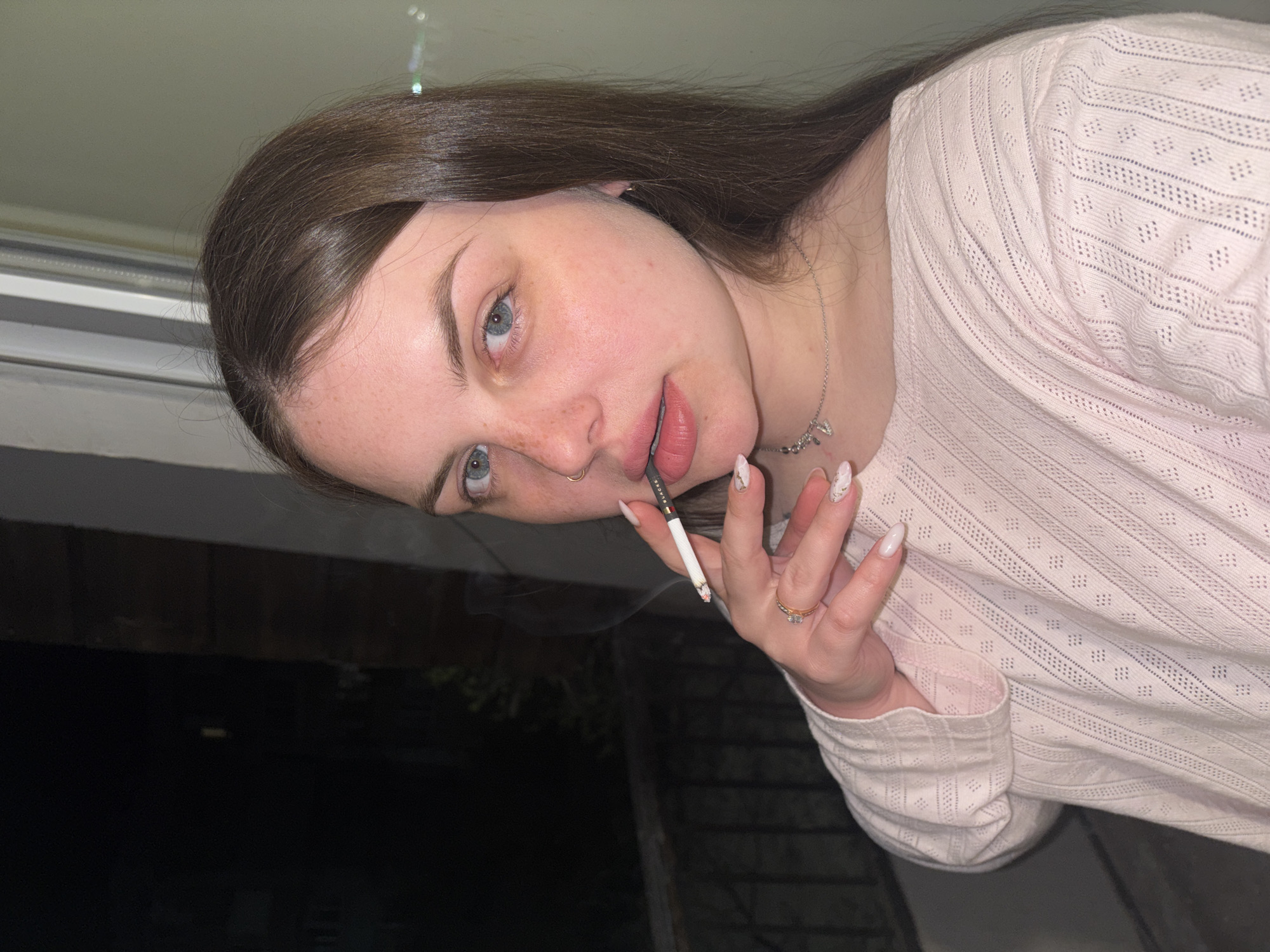 A young woman with long, straight dark brown hair and light blue eyes is depicted in a close-up, angled shot. Her fair skin shows a scattering of freckles across her nose and cheeks. She wears a light pink, textured long-sleeved top with an eyelet pattern. A silver necklace with a small charm hangs around her neck, and she has a silver hoop piercing in her right nostril. On her left ring finger, she wears a ring with two stones, and her fingernails are long with a white marbled design.
The woman is smoking a cigarette, which is held between her lips by her left hand. The brand "FUCHS" is visible on the cigarette filter, and a wisp of smoke is present. She looks directly into the camera with a neutral expression. The setting appears to be outdoors at night, indicated by the dark, indistinct background, which includes hints of a window frame and possibly distant structures or foliage. The location is Chornomorskoe, Ukraine.