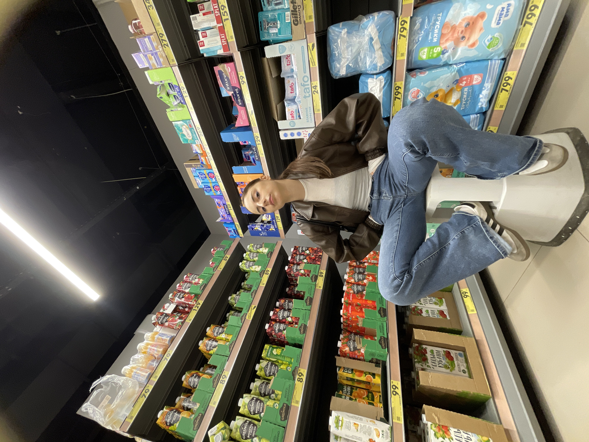 A person sits on a stepping stool in a store aisle filled with products, likely in Moscow, Russia. The individual, wearing a brown jacket and jeans, looks at the camera. The aisles are stocked with fruit juice, personal hygiene products, and diapers, each labeled with prices in rubles.