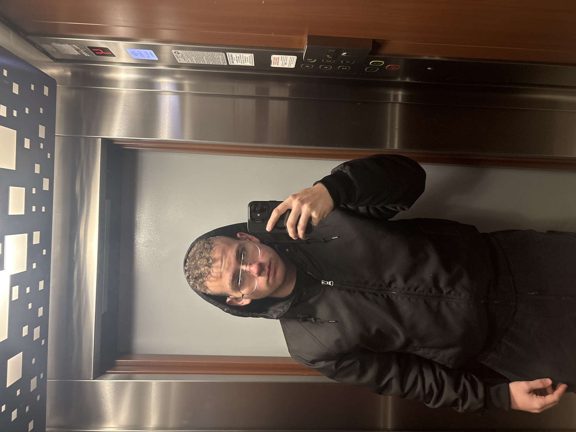 A young adult is taking a selfie in an elevator. The elevator has brushed metal walls, a dark wooden trim, and a control panel with numbered buttons. The floor indicator shows "2" in red. The individual is wearing a black hooded jacket and glasses. The location is unspecified but it is not Plovdiv, Bulgaria.