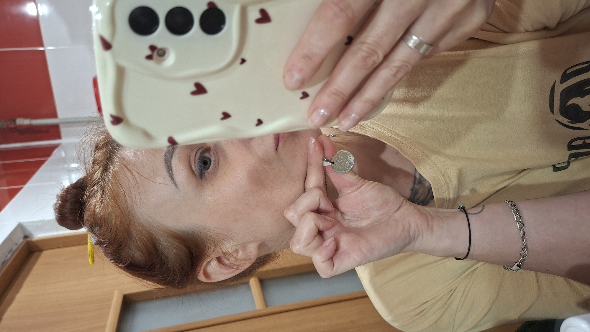 A woman with reddish-brown hair pulled into a bun on top of her head is holding her smartphone, which has a cream-colored case with small red hearts, in her right hand to take a selfie. She is wearing a tan t-shirt and a silver pendant necklace. Her left hand is holding the pendant. She is also wearing a thin black bracelet on her left wrist and a silver chain bracelet on her right wrist. A silver ring is on her right ring finger. Her hair is held up with a yellow hair clip. The background appears to be a bathroom with tiled walls, one of which is red and white. There is a wooden door on the left side of the image. The scene is indoors.