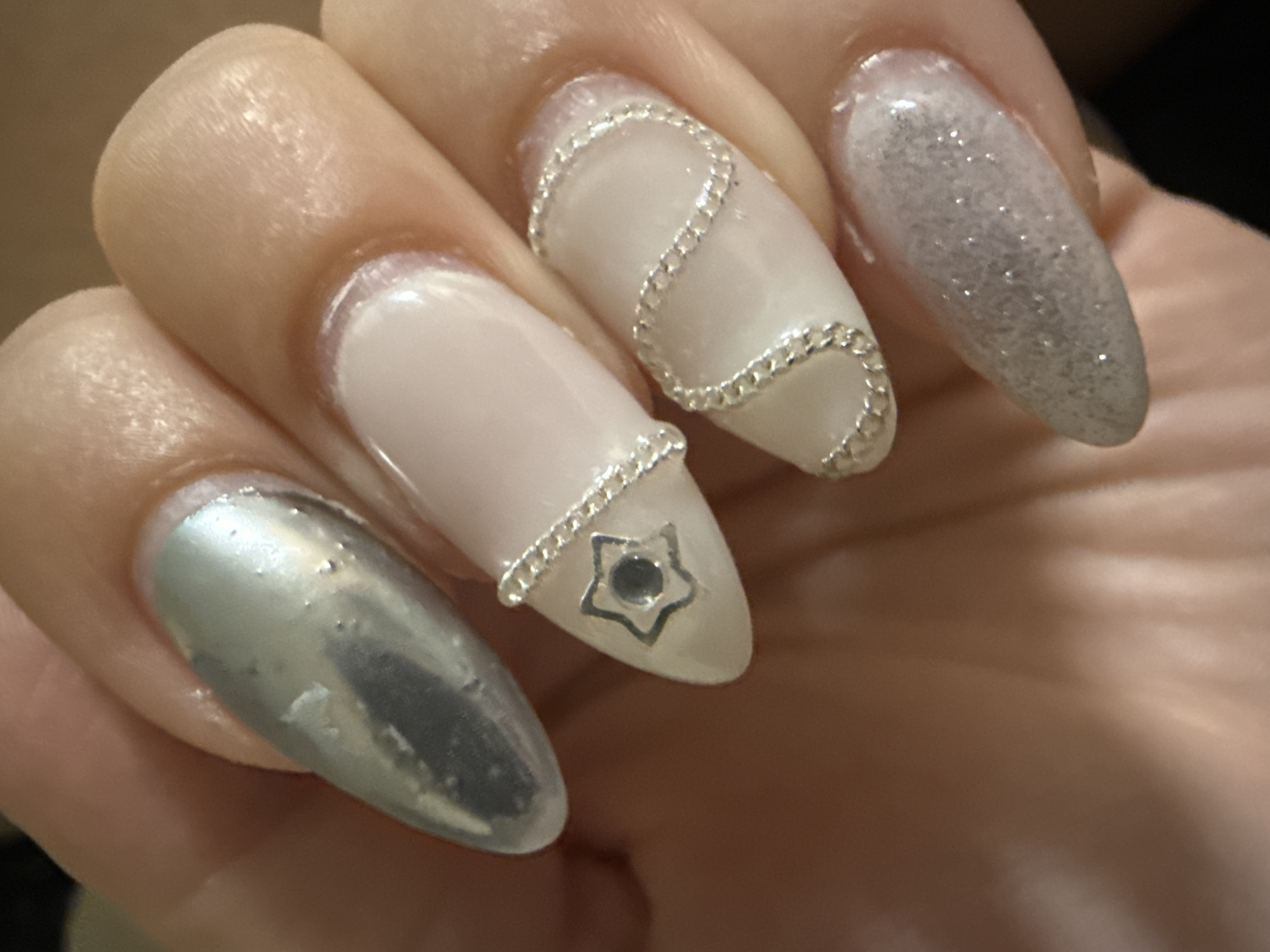 The image shows a close-up of a hand with four extended fingernails. The fingernails have been decorated with different designs: one has a silver, reflective, metallic coating; another has a pale, opaque base with a silver chain wrapped in a wavy pattern; another has a pale, opaque base with a silver chain band near the tip and a silver star-shaped stud; and the final one has a pale base with silver glitter. The background is blurred and indistinct, but appears to be composed of skin tones.