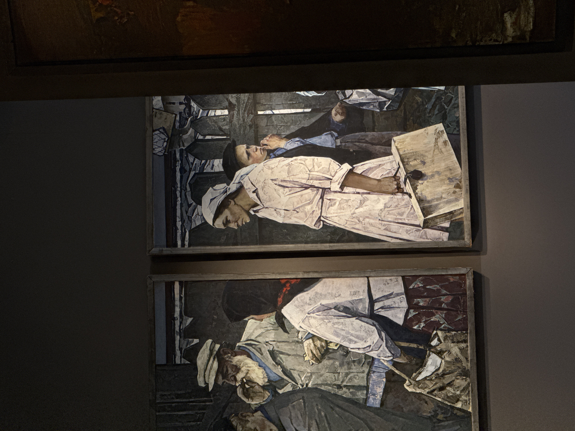 Two framed paintings are displayed on a wall. The top painting depicts two figures lying down, with one in a light-colored outfit and the other in darker clothing. The bottom painting also features figures lying down, with one appearing older and bearded. Both paintings have a similar style. The location is likely a museum or gallery in Moscow, Russia, given the presence of artworks on display.