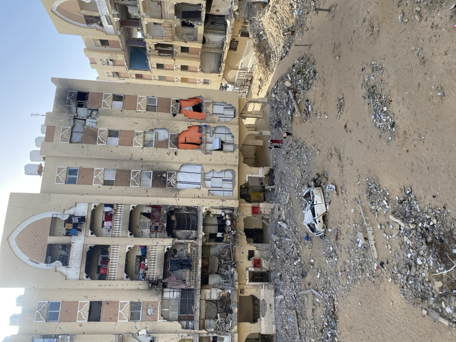 This image captures a scene of widespread destruction in the city of Khan Yunis, Palestinian Territory, under a clear, light blue sky. The central feature is a multi-story building, likely residential, constructed of light-colored material with ornate arched motifs above windows and balconies. The building exhibits extensive damage, including numerous small caliber impact holes peppered across its facade, shattered windows, and considerable structural compromise. Several balconies are damaged, with clothing items such as red shirts, blue shirts, and dark garments still hanging on lines. The fifth floor on the right side of the main building shows significant charring and collapsed sections, indicating fire damage. Lower-level openings on the right side are partially covered by large orange tarpaulins. To the right of the main structure, a second, similarly styled building is partially visible, also displaying substantial damage, exposed interiors, and collapsed sections.
The ground area in the foreground and mid-ground is a debris field comprising sand mixed with large quantities of rubble, concrete fragments, and twisted metal. A severely damaged, white four-door sedan, crushed and distorted, rests amidst the rubble in the lower-left-center. Two individuals are visible near the base of the main building: one person wears a red top and dark pants and stands upright, while another person in dark clothing appears partially obscured or bent over. A third individual in dark clothing is further to the right.