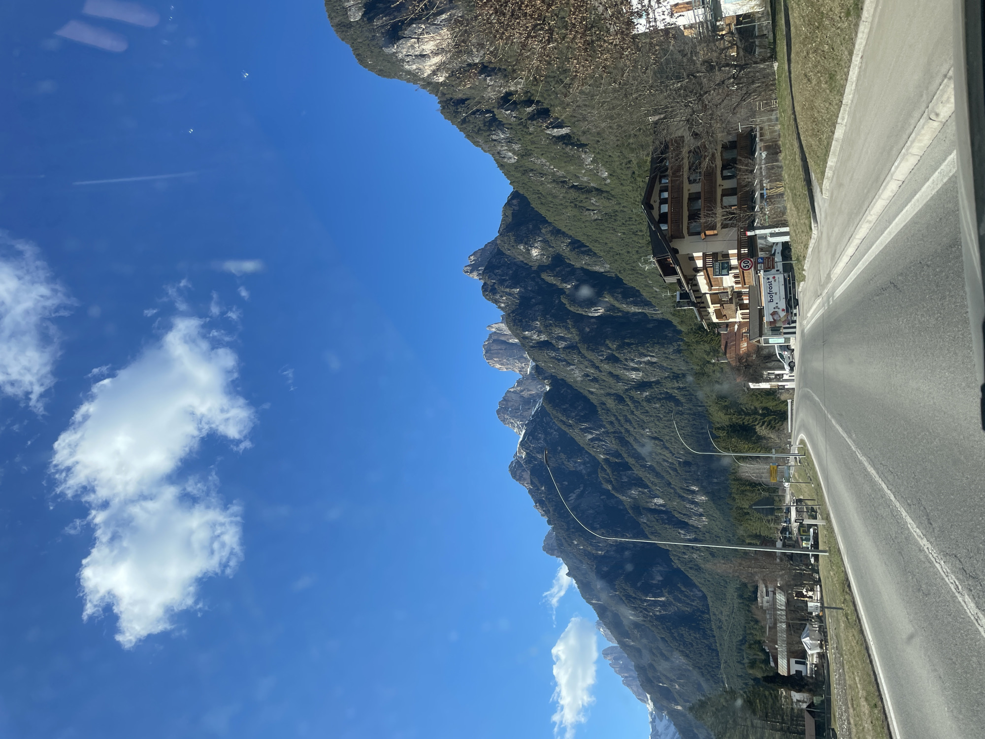 Auronzo di Cadore, Italy: A slightly angled shot shows a road curving to the right, bordered by green grass and a building to its right. Light posts line the road. To the left, a mountain range rises sharply, with some bare rock and trees near its peak. The sky is a clear blue with scattered white clouds. A building with the name "bofrost" can be seen.