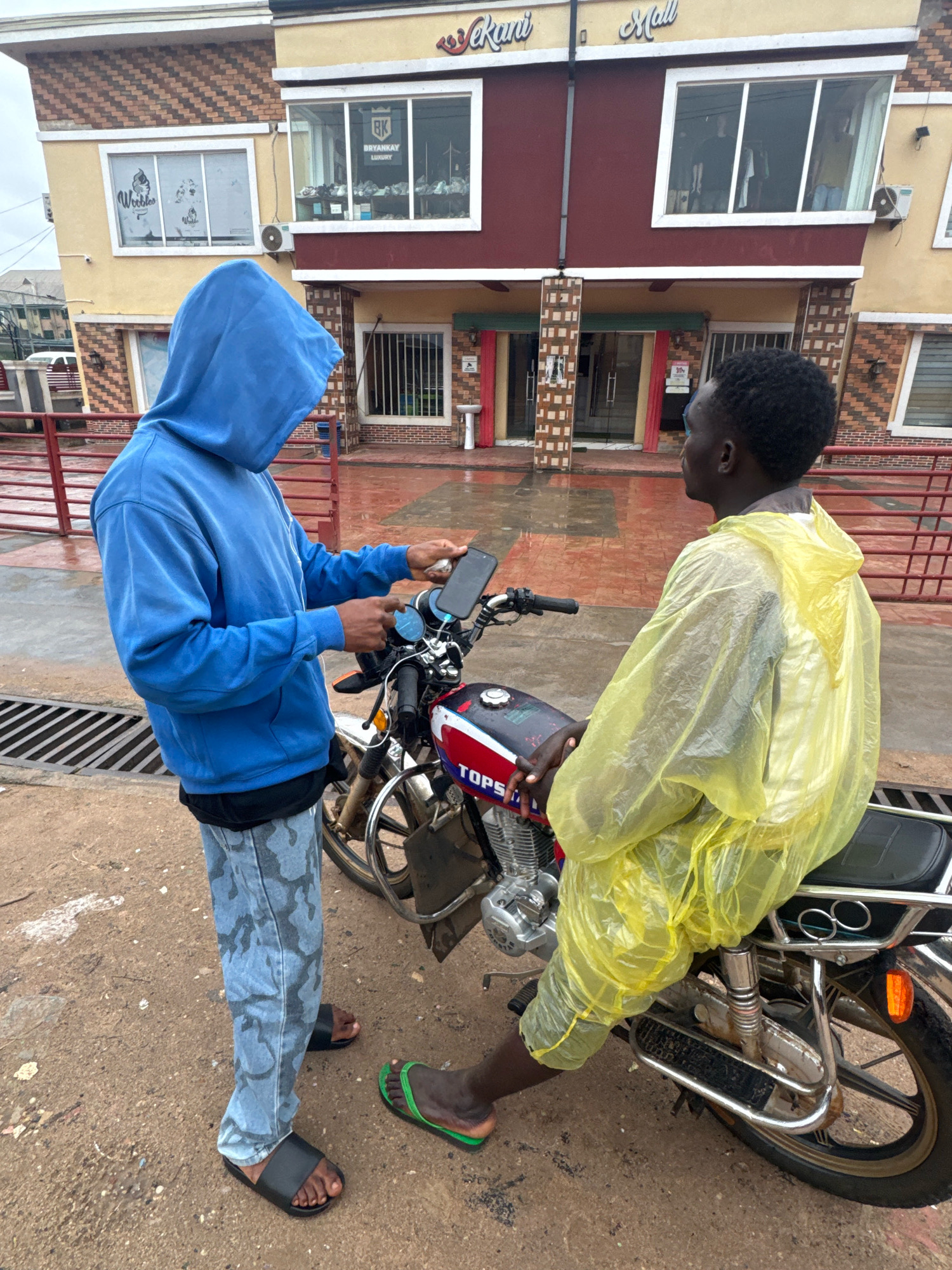 In Ihiagwa, Nigeria, on a cloudy day, two young Black males stand near a red and white motorcycle parked on a wet, paved ground. The male on the left, wearing a blue hooded sweatshirt and camouflage-patterned pants, holds a smartphone in his right hand. The male on the right, seated on the motorcycle, is dressed in a bright yellow disposable rain poncho. Behind them stands a two-story building with brown and yellow facades, featuring windows and signage indicating "Vekani Mall." The lower level of the building has covered entrances, and there are red metal railings along the sidewalk. The ground is damp, suggesting recent rain.