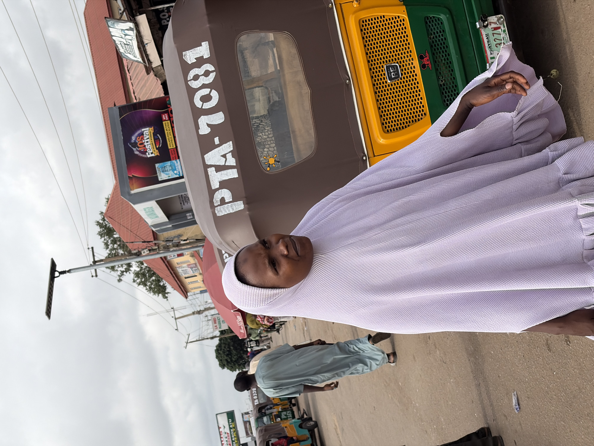 A medium shot captures a person in a light purple textured garment and head covering, standing beside a motorized tricycle (keke napep) on a street in Gura Top, Nigeria. The person looks directly at the camera with a neutral expression.
The tricycle, visible in green, yellow, and brown, has "PTA-7081" painted in white on its brown canopy. Its front grille features "RE" branding. A Nigerian license plate, partly visible, shows "PLATEAU" and "27Z77", identifying its origin as Plateau State.
In the background, another person in a light blue garment walks further down the street. Multiple other tricycles are partially visible. The scene is set on a paved street with commercial buildings lining it. One building has a "JOS Festival" banner advertising an event in "RAYFIELD JOS, PLATEAU" and mentioning "PHILADELPHIA". Another sign indicates "LA MED PHARMACY LTD". A street lamp with a solar panel stands on the left. The sky is overcast. The overall impression is a bustling street scene in a Nigerian town or city.