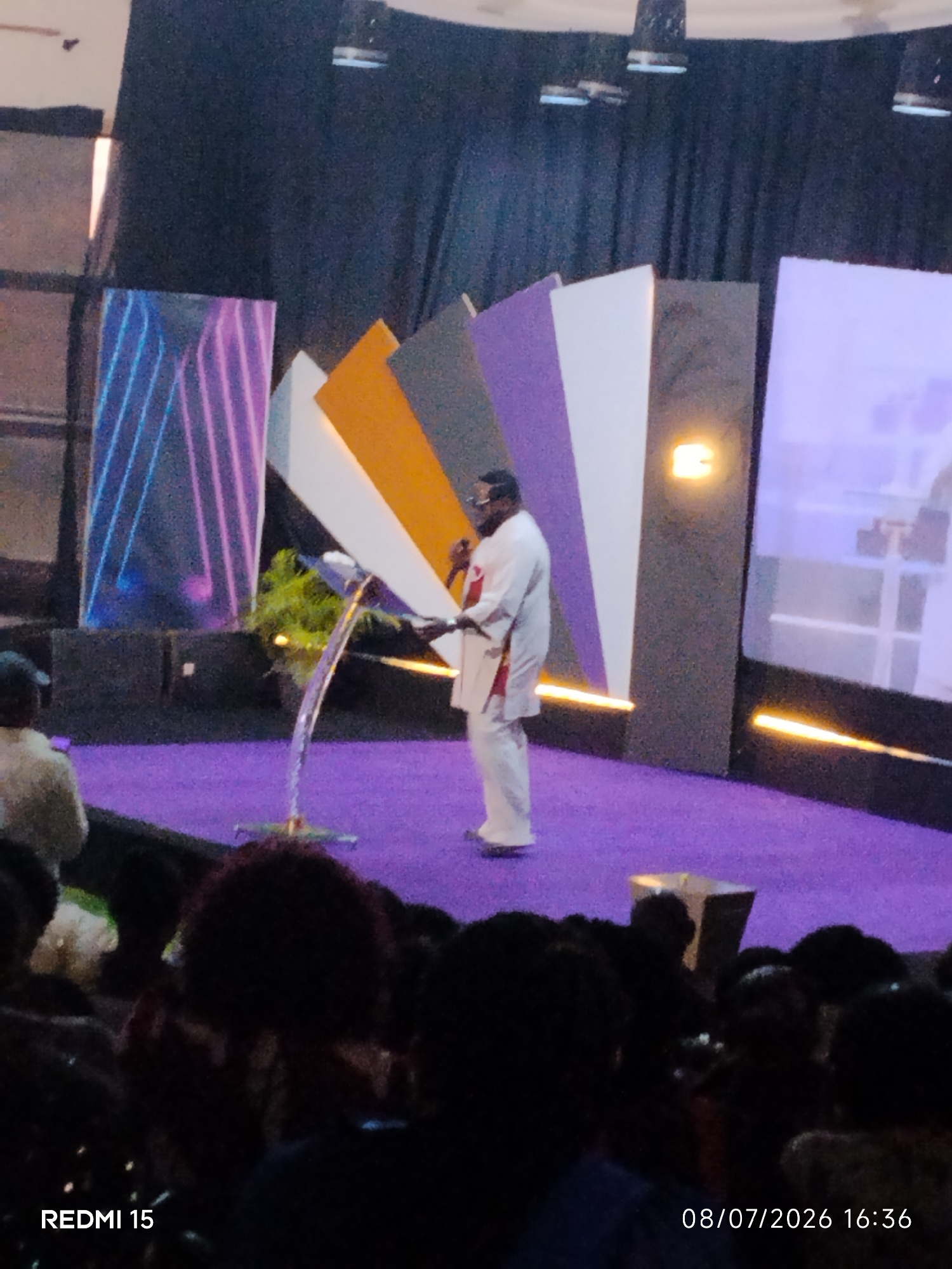 A man in a white robe stands at a podium on a purple carpeted stage, addressing an unseen audience. He holds a microphone in his right hand. The stage is decorated with abstract, colorful panels and has bright yellow lighting strips. In the foreground, the backs of several audience members are visible, mostly in dark clothing. The image includes a watermark in the bottom left corner that reads "REDMI 15" and in the bottom right corner the date and time "08/07/2026 16:36". While the image is undated, based on the watermark and typical event settings, this scene is likely from a church service or conference in Aba, Nigeria, given the presence of a prominent religious leader.