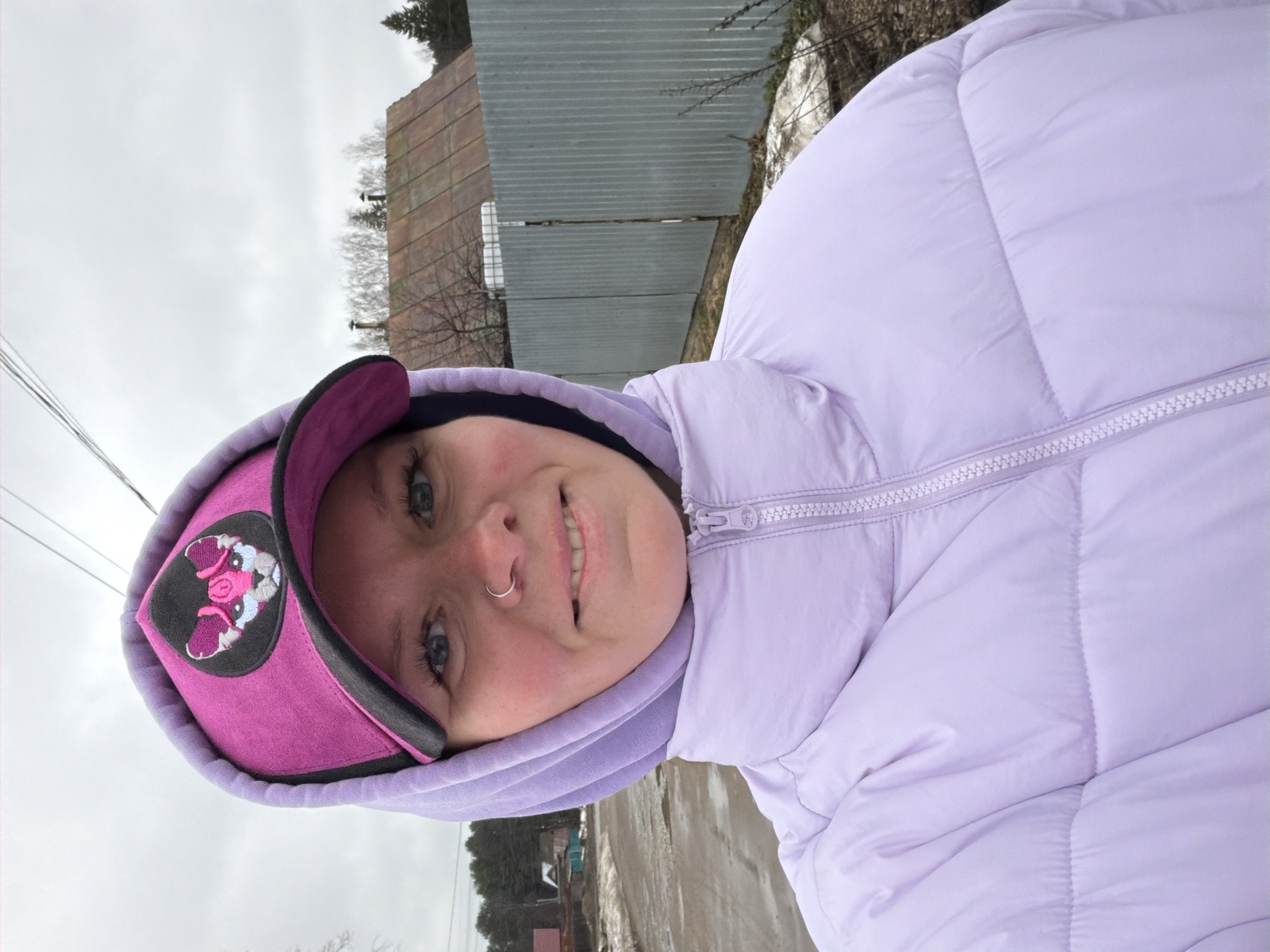 A young woman is facing the camera with a slight smile. She is wearing a light purple puffer jacket with a hood that is pulled up. She also has on a bright pink cap with a black brim and a cartoon patch on the side. The woman has light skin, blue eyes, and a nose ring. The background features a cloudy sky, a corrugated metal fence, bare trees, and a small building. The location is likely Chaikovsky, Russia, based on context cues like architecture and environment typical of Russian small towns. The ground appears to be damp or possibly snow-covered.