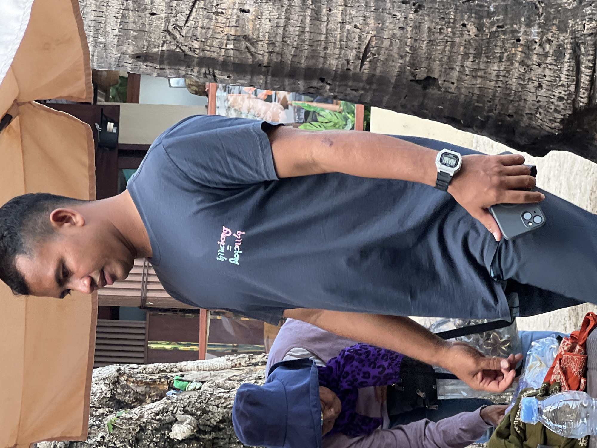 In Kuta, Indonesia, a man stands on a street next to a large palm tree. He wears a dark grey t-shirt with a colorful logo and dark pants. On his left wrist is a white digital watch. He holds a grey smartphone in his right hand, near his hip. To the man's left, another person wearing a purple scarf and a blue hat is partially visible, interacting with items laid out on a surface in front of them. A clear plastic bottle of water is also visible. The background shows an outdoor market setting with a striped awning and wooden structures.