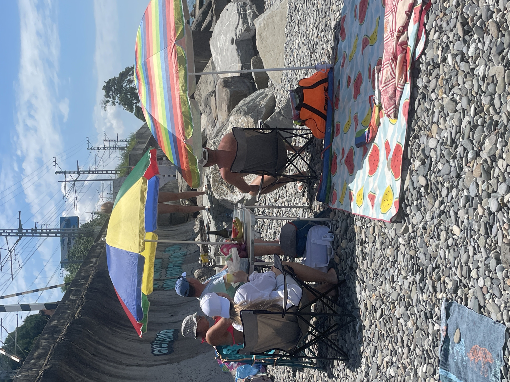 The image captures a sunny day at a pebbled beach in Sochi, Russia, with several people enjoying a relaxed outing. The scene is set against a backdrop of large rocks and a bright blue sky with scattered clouds, suggesting pleasant weather.
In the foreground, a group of individuals are seated in foldable camping chairs under colorful beach umbrellas. One umbrella is a vibrant rainbow pattern, and another features segments of yellow, blue, green, and red. A patterned beach blanket with watermelon and banana designs is spread on the pebbles. A person wearing a white cap and a pink bikini is visible in the lower right quadrant, facing away from the camera. Nearby, another person in a white shirt and white cap is seated, and a third person in a blue t-shirt and blue cap is also present. A small table is set up with a bottle of wine and some drinks, indicating a picnic or gathering.
Further back, another person sits in a chair under the rainbow umbrella, wearing goggles around their neck and an orange life vest with the word "PRIDE" printed on it. The background reveals more large rocks and the presence of railway tracks and overhead power lines, suggesting the beach is situated near a transportation route. Graffiti is visible on a concrete embankment to the left, adding a touch of urban detail to the natural setting. The overall mood appears to be one of leisure and enjoyment on a warm day by the sea.